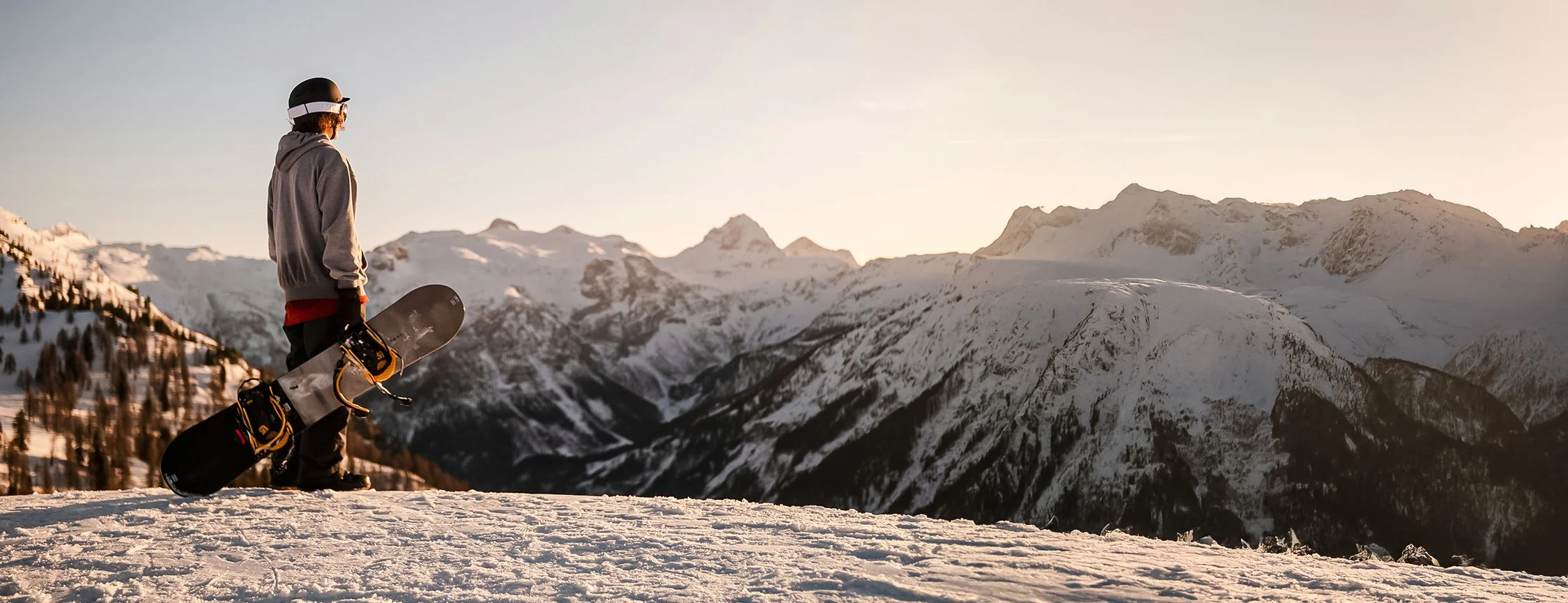 Skier with snowboard standing on snowy mountain, overlooking snow-covered mountain range at sunset.