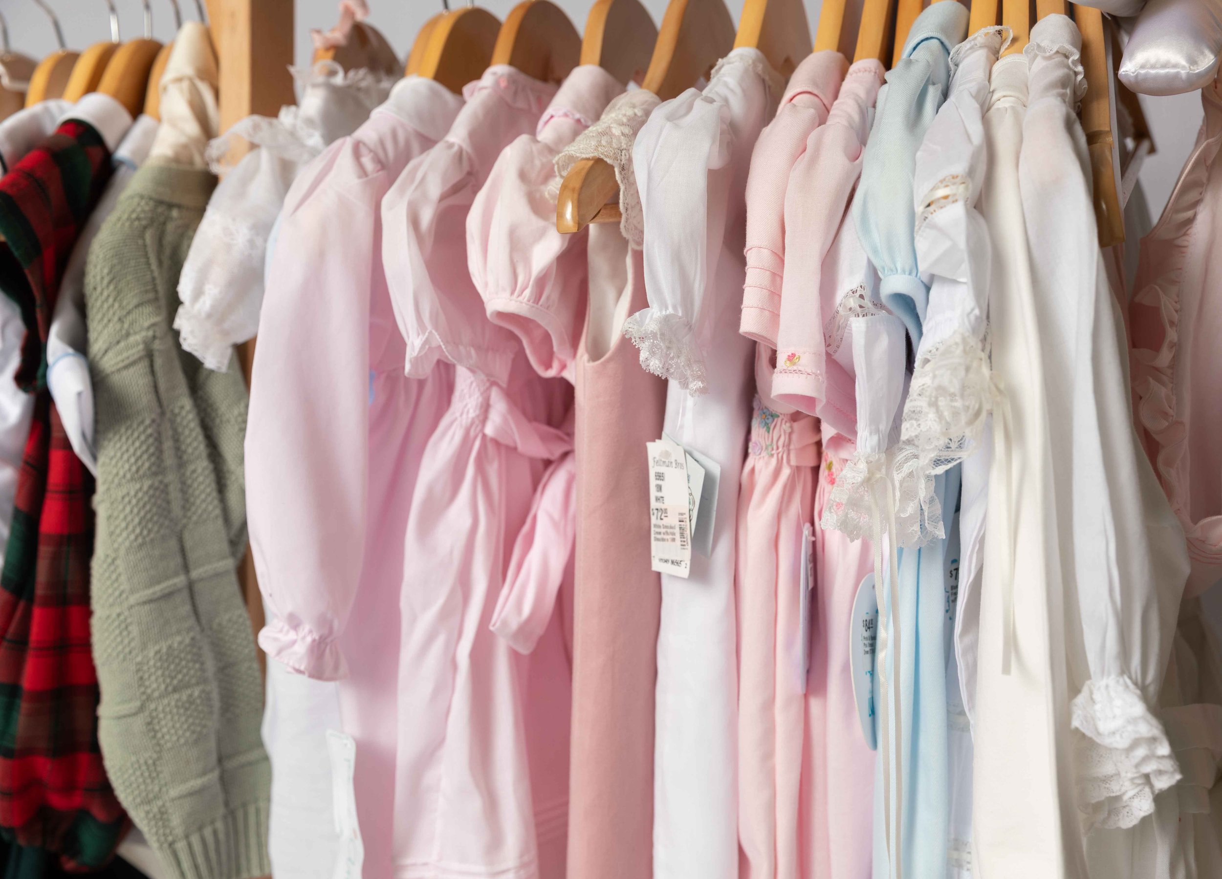 Various children's clothing items hanging on a rack, including white, beige, and pink dresses and shirts.