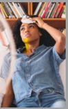 A young woman wearing a blue denim shirt and yellow scarf looks upwards, sitting in front of bookshelves.