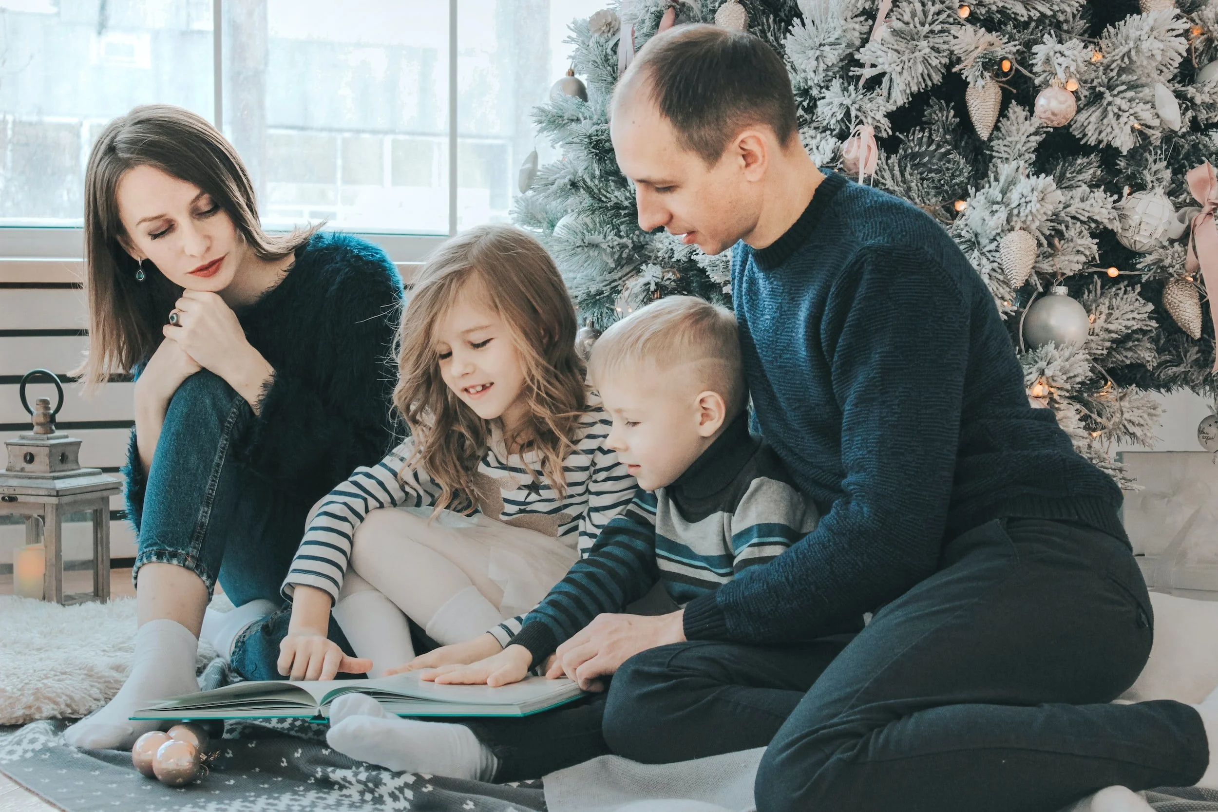A family of four reading a book together in front of a decorated Christmas tree.
