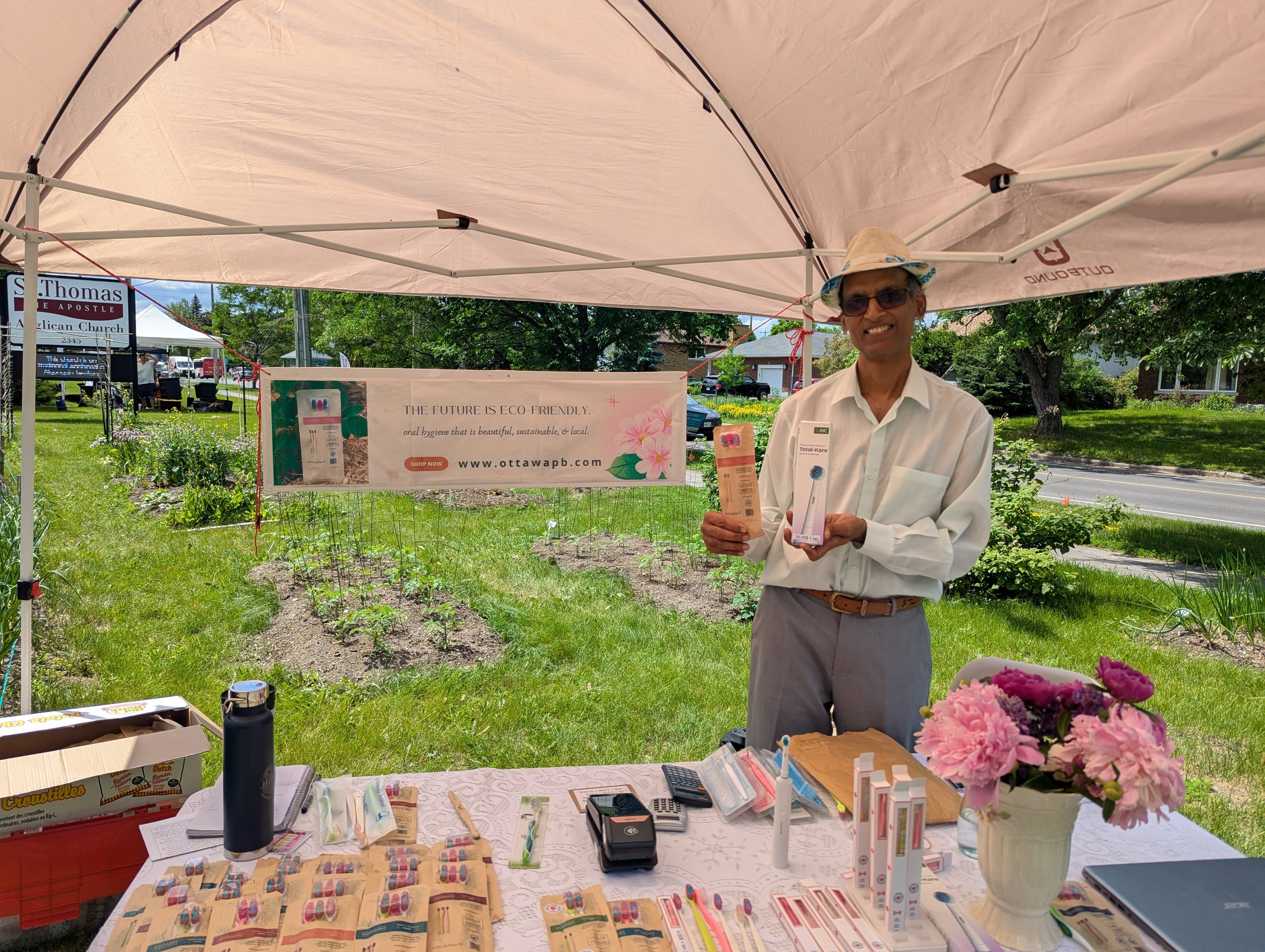 A man wearing glasses and a hat smiling while holding two dental product packages at an outdoor booth under a canopy. The booth displays dental supplies and a banner with ecological and local dental hygiene message. There are flowers on the table and