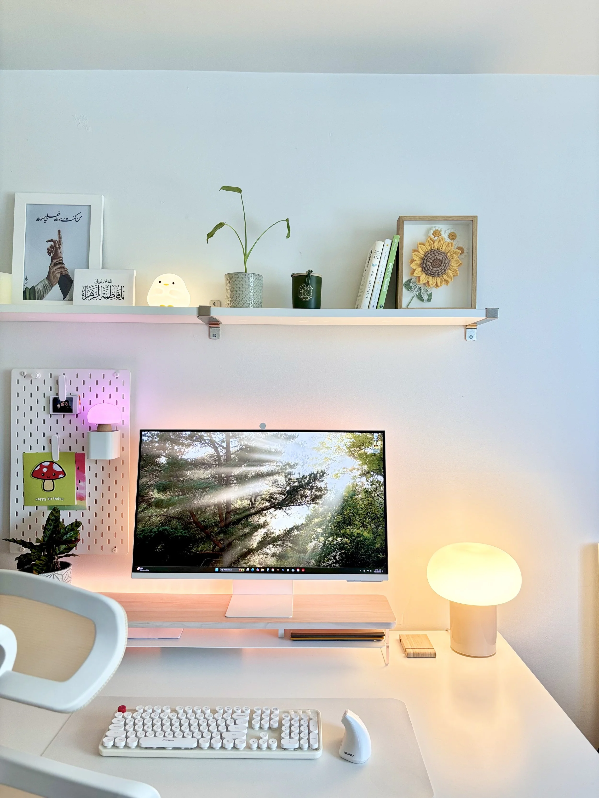 A tidy home office with a computer monitor, keyboard, and mouse on a white desk. There is a small lamp with a rounded shade, a potted plant, and a pegboard with small items on the left side. A wood shelf above holds books, framed art, a small framed sunflower, and a decorative light.