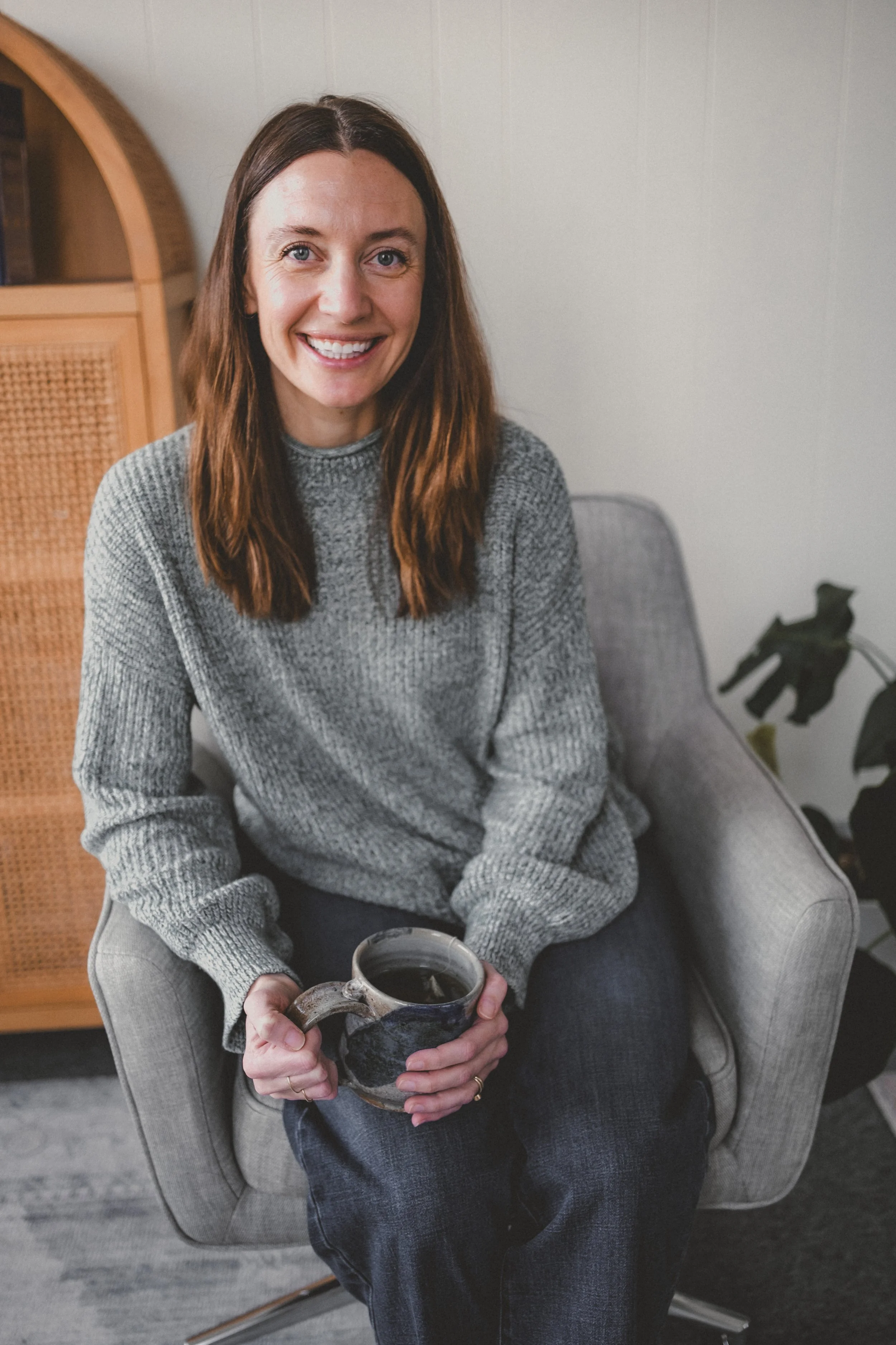 A woman with shoulder-length brown hair, wearing a gray knit sweater, sitting in a light gray armchair, holding a dark ceramic mug, smiling at the camera, in a cozy indoor setting with a wooden cabinet and a plant in the background.