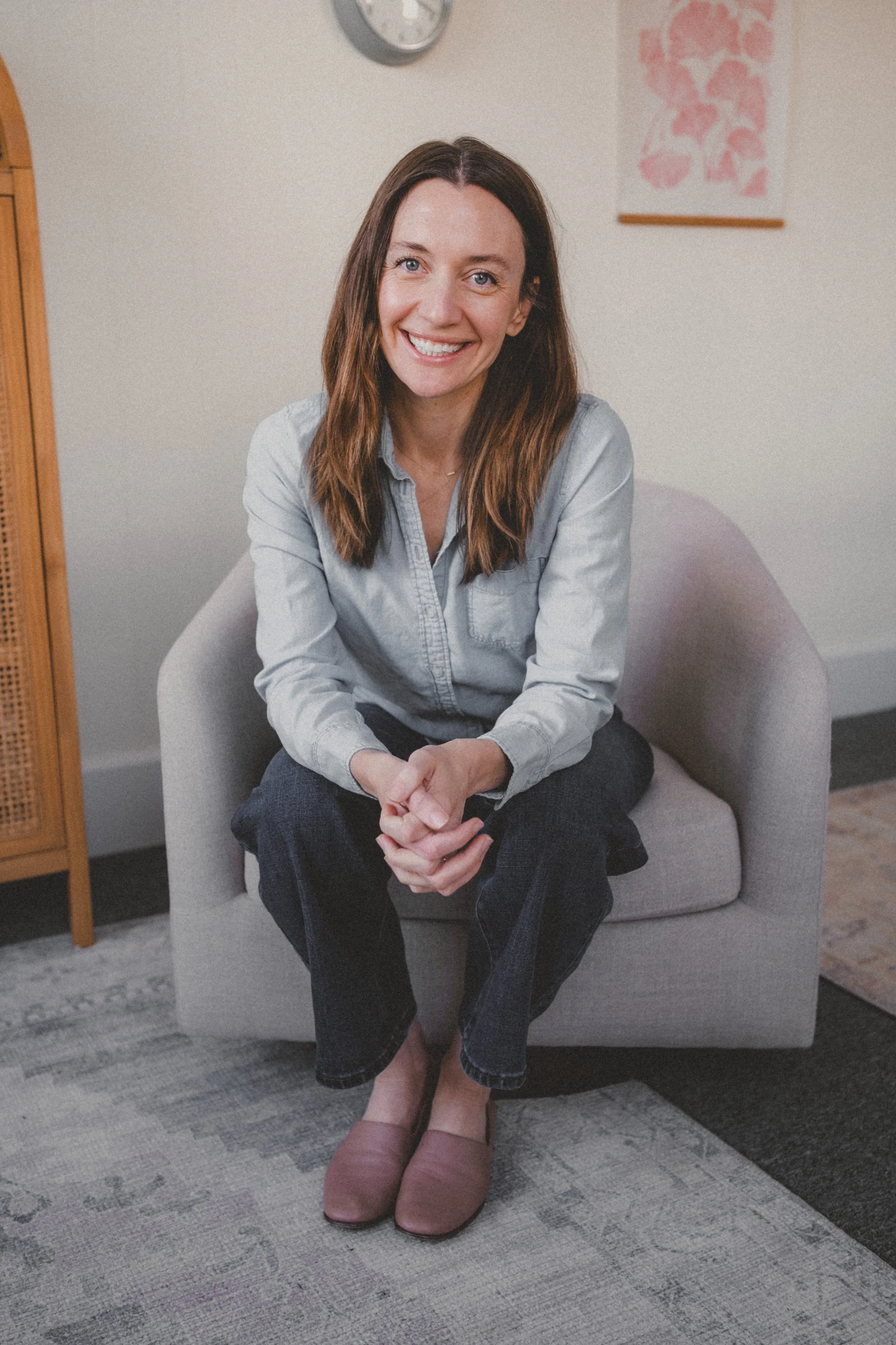 A woman with long brown hair, wearing a light blue button-down shirt, dark jeans, and pink slip-on shoes, sitting on a gray armchair in a room with minimal decor, smiling at the camera.