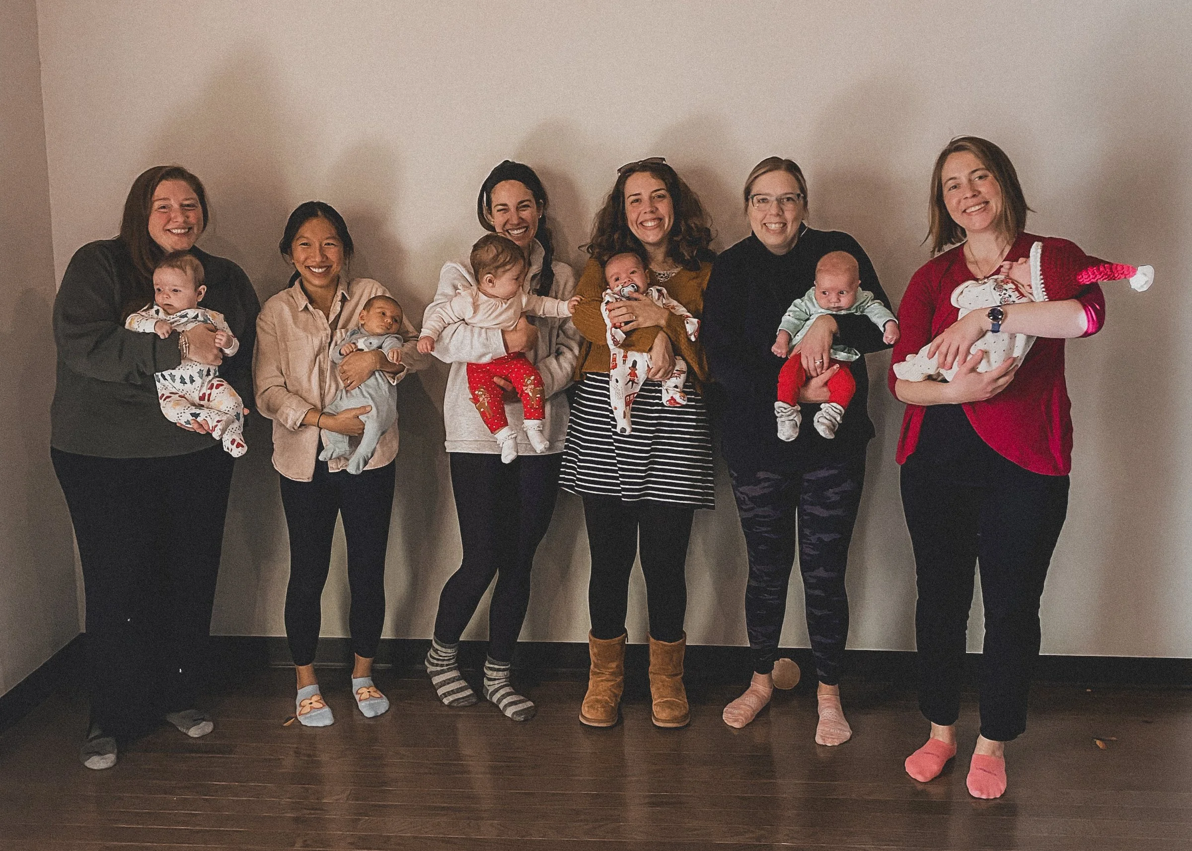 Seven women standing in a row against a plain wall, each holding a baby, smiling, some dressed in casual and some in festive holiday attire.