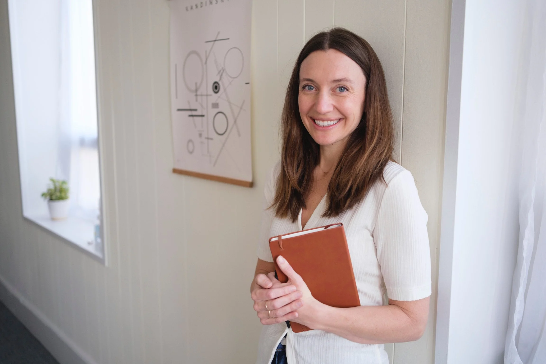 A woman with long brown hair, smiling and holding a brown notebook, standing in a well-lit room with white walls and a window with white curtains. There is a small potted plant on the windowsill and abstract artwork on the wall behind her.