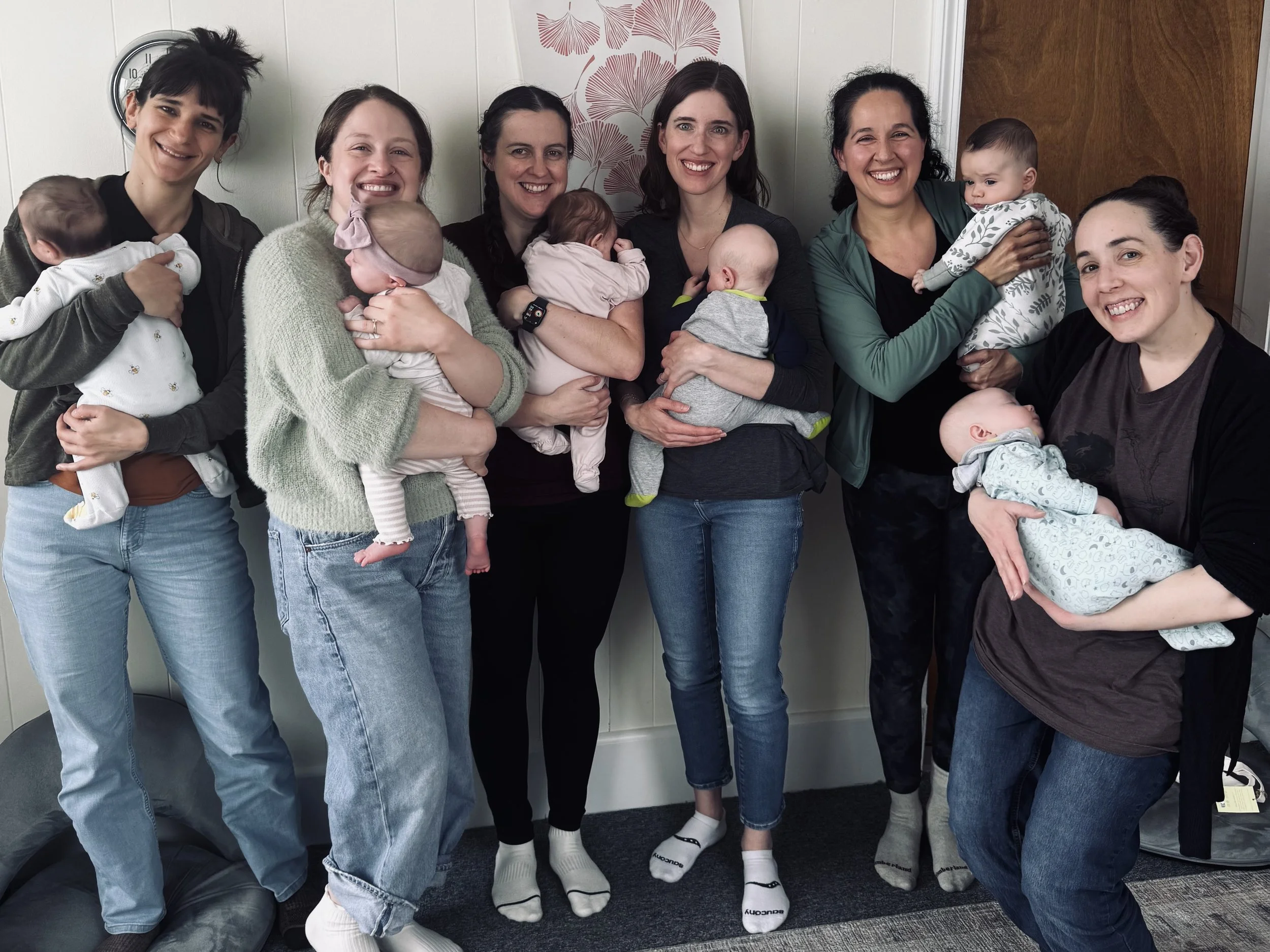 Group of women holding babies, smiling and standing together indoors.