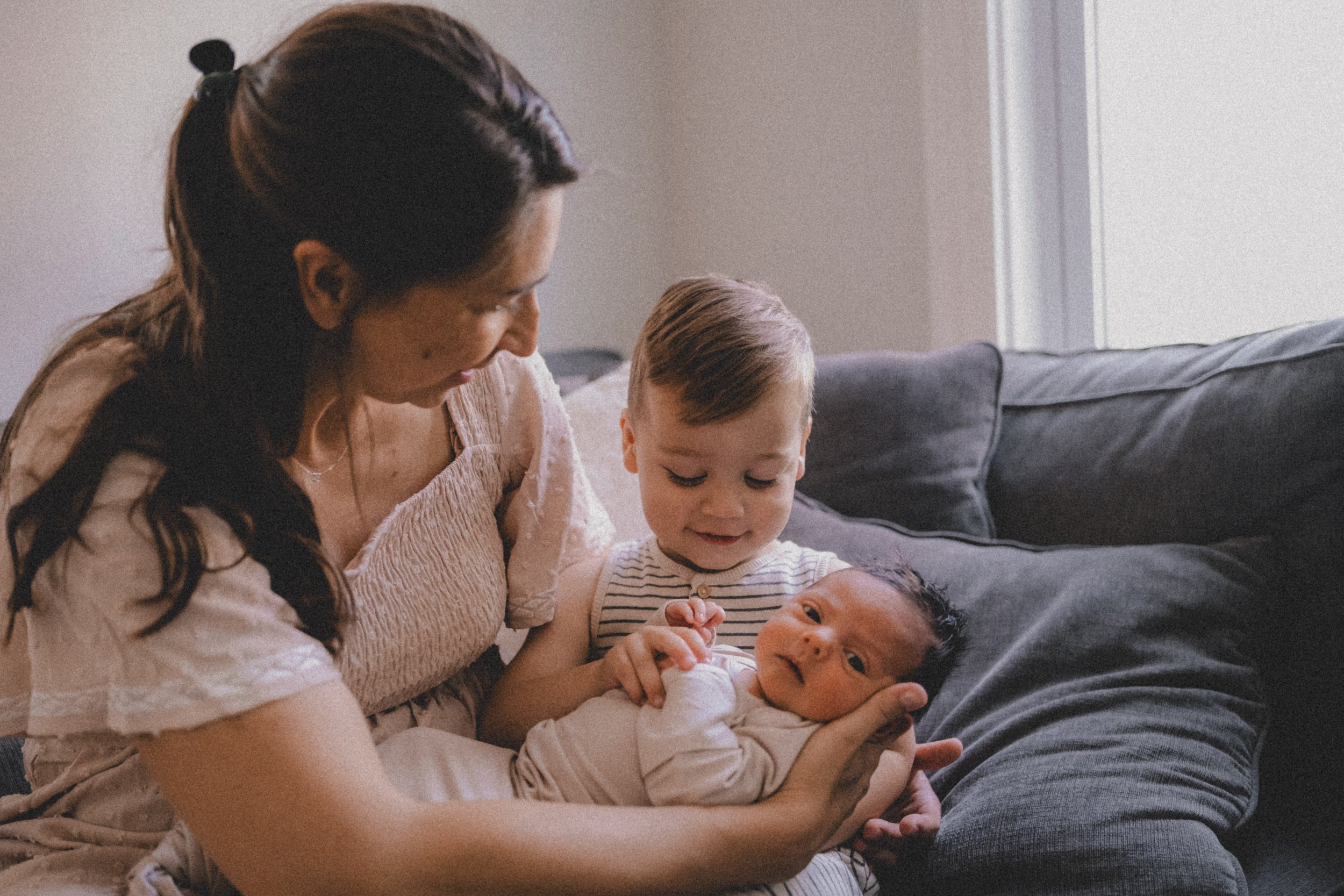 A woman with dark hair holding a newborn baby on her lap, a young boy with blond hair looking at the baby, all sitting on a dark gray couch in a cozy room with soft lighting.