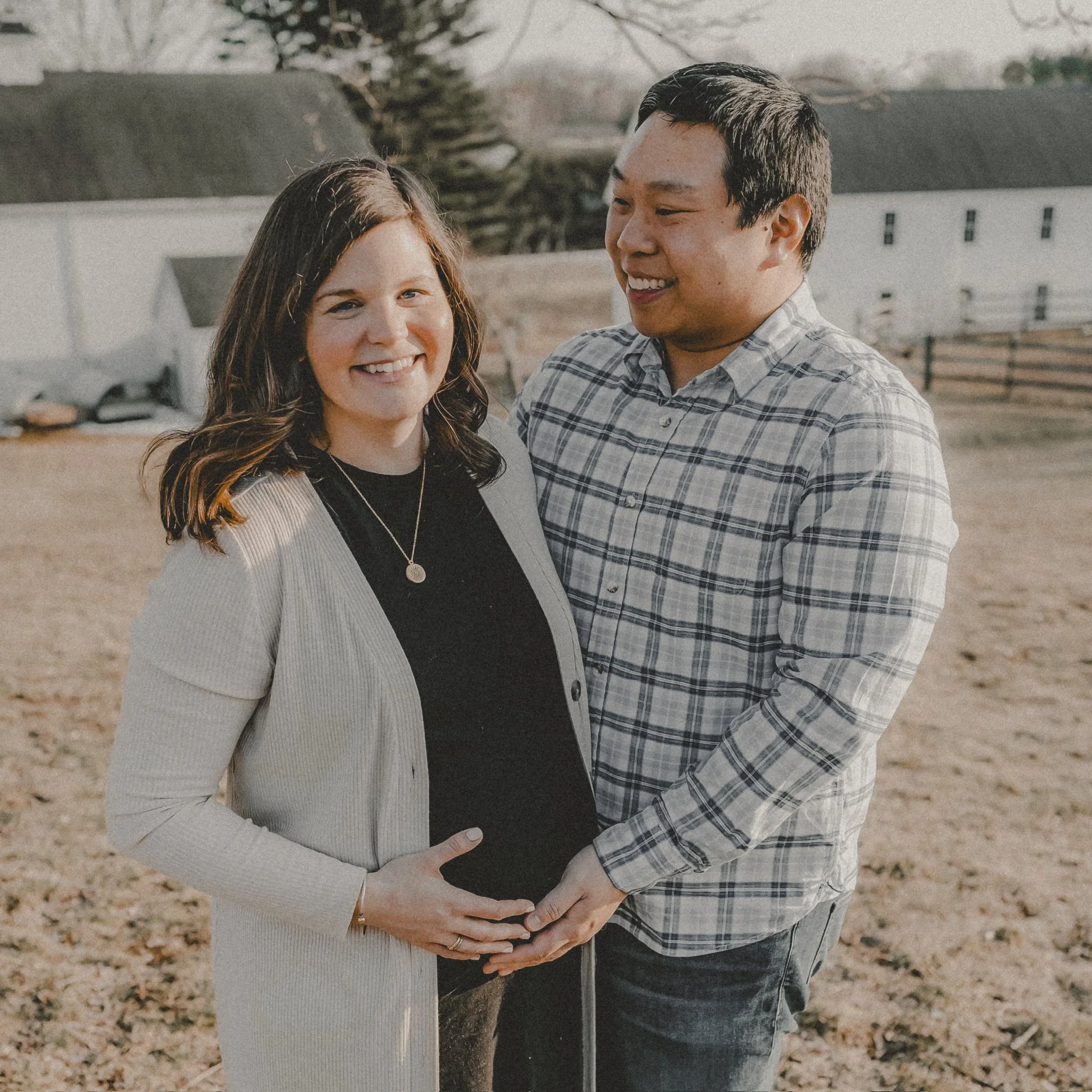 A smiling pregnant woman with long brown hair wearing a black top and light cardigan, holding her baby bump, stands outdoors with a man who has short dark hair, wearing a plaid shirt, with their hands on her belly, in a rural setting with barns and trees in the background.