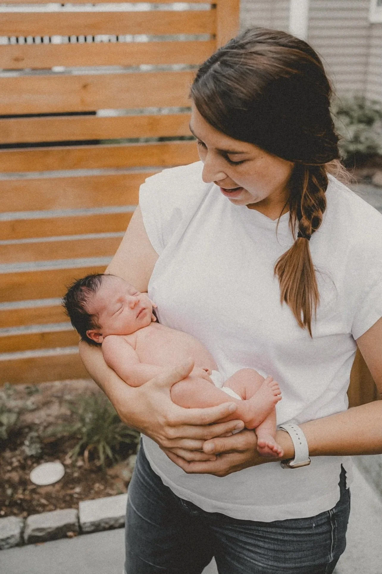 A woman with long braided hair holding a newborn baby outdoors in front of a wooden fence.