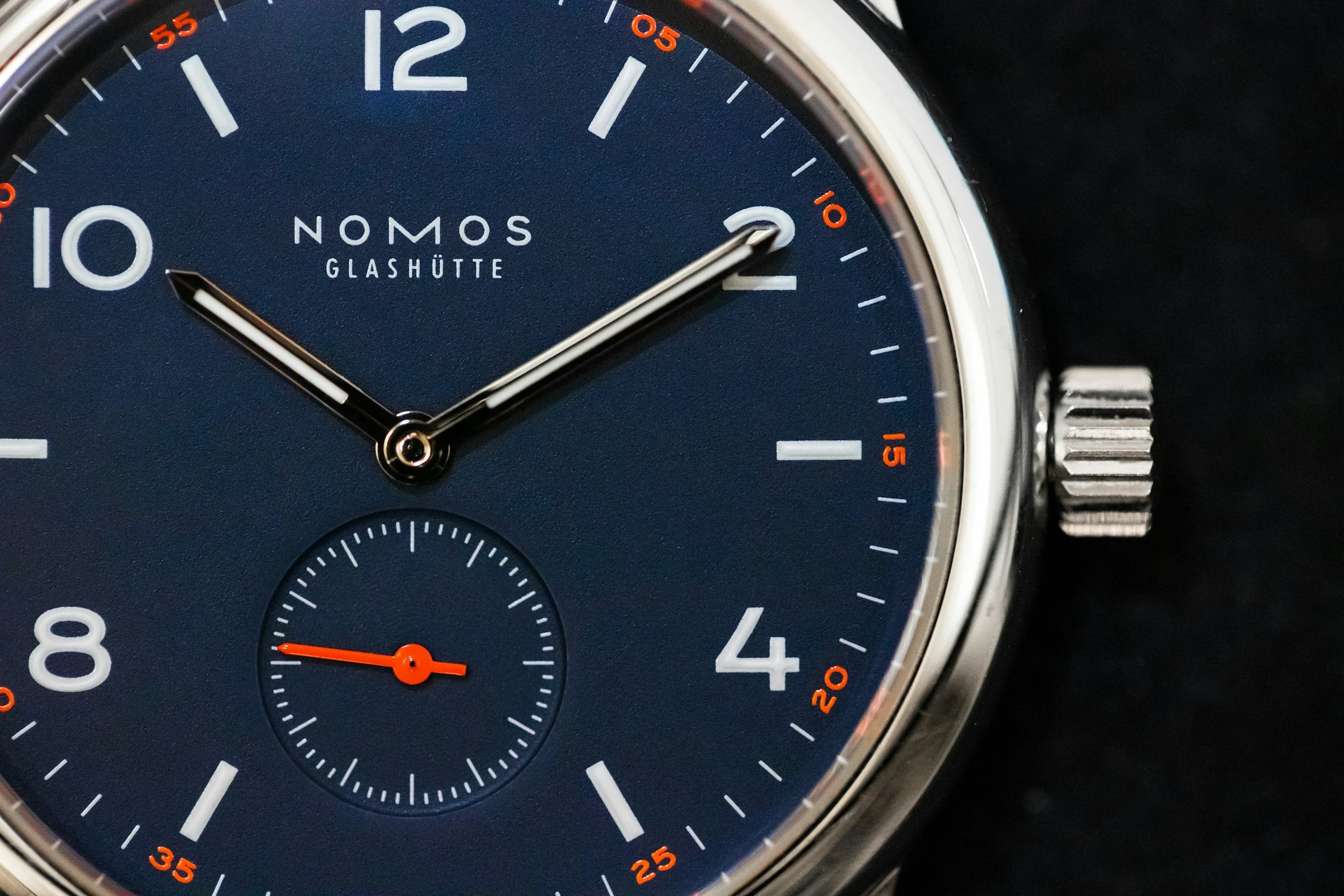 Close-up of a Nomos Glashütte wristwatch with a navy blue dial, silver watch hands, and a smaller seconds subdial at the 6 o'clock position, on a metal case with a ridged crown.