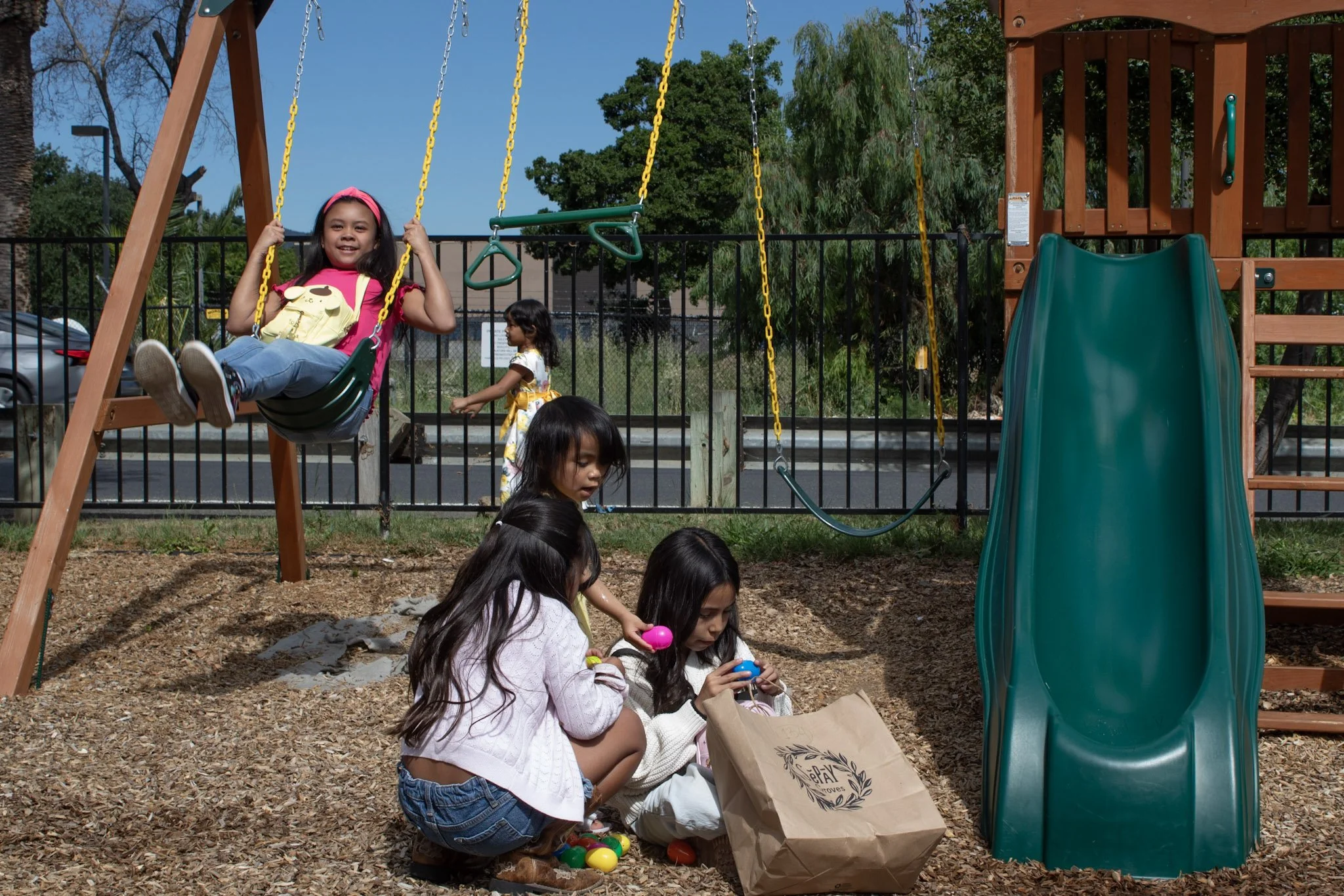 Children playing on a playground, with a girl on a swing smiling and holding the chains, other kids sitting on the wood chip ground playing with colorful eggs, a slide, and a park fence in the background.