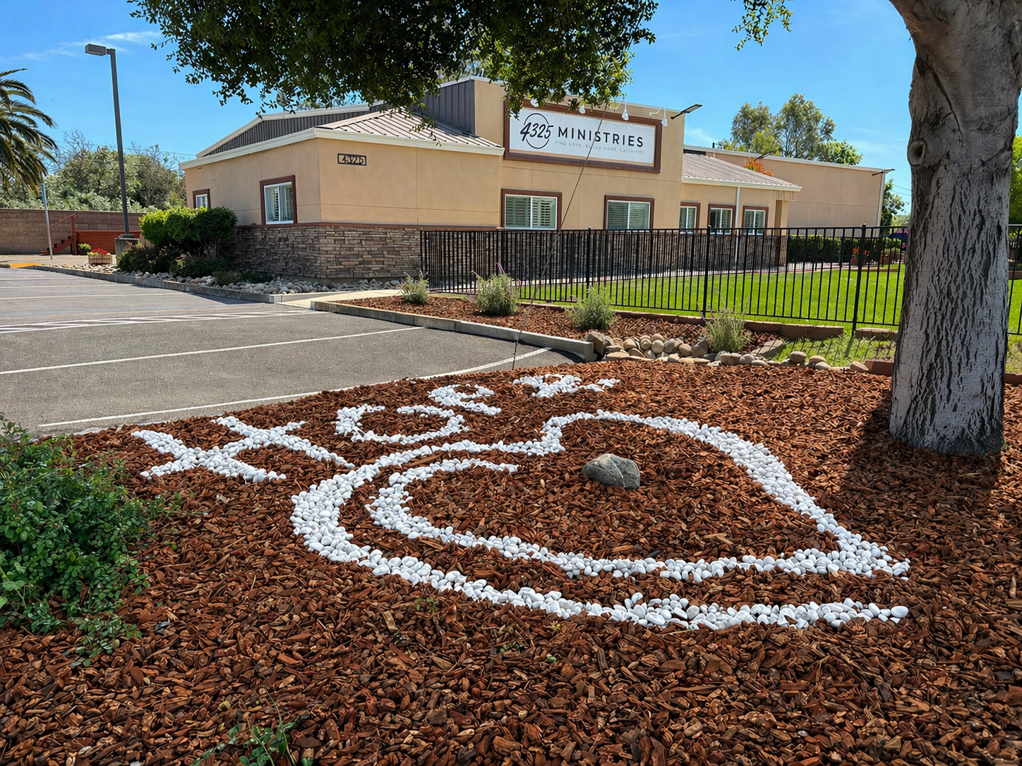 Outside view of a beige building with a sign that reads '4325 Ministries.' In the foreground, there is a landscaped area with brown mulch, white rocks spelling out a word, and a tree on the right. There is a parking lot with a few empty spaces in front, and a small grassy area with bushes and shrubs.