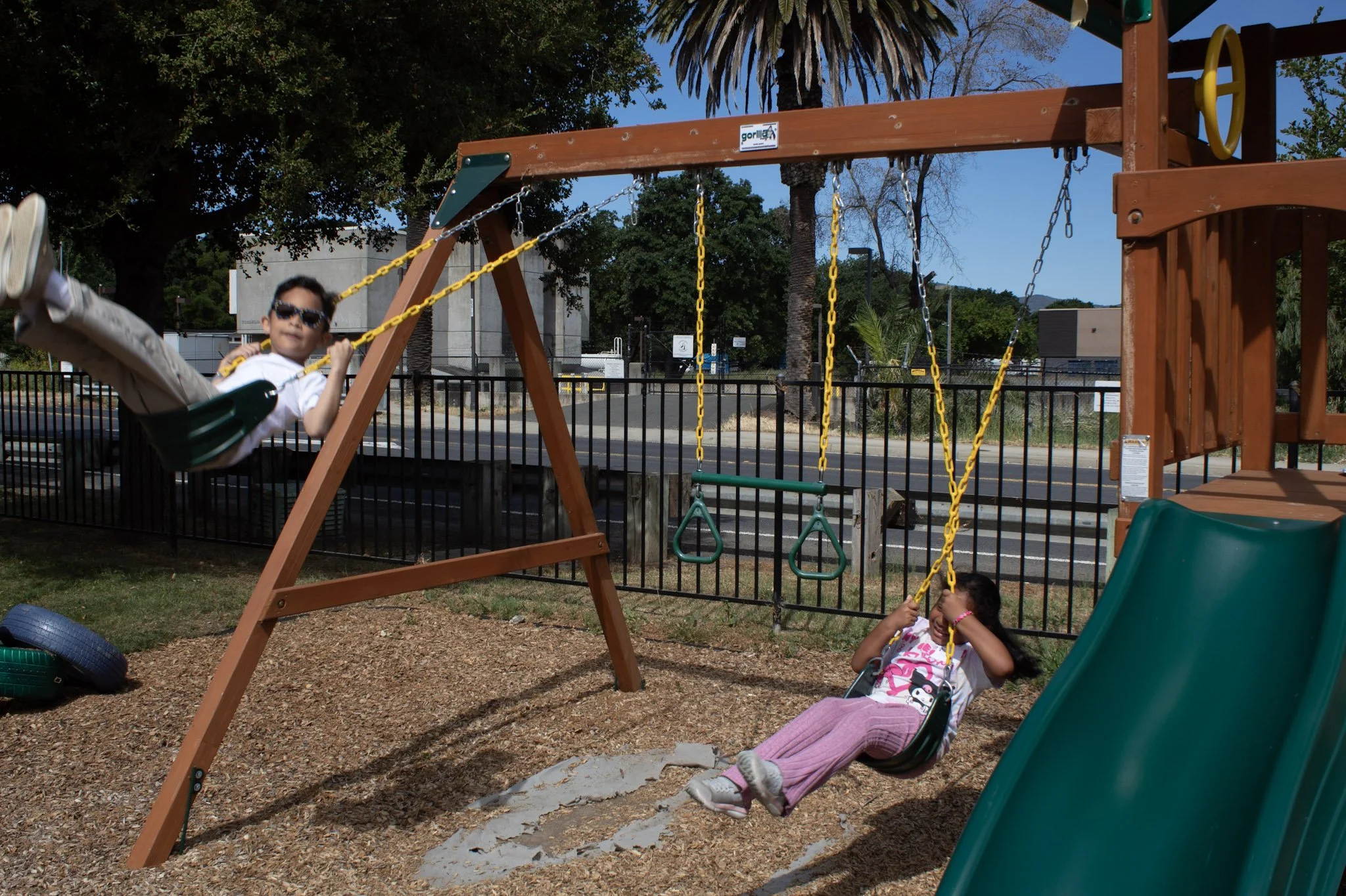 Children playing on swings at a playground with trees and a fence in the background.
