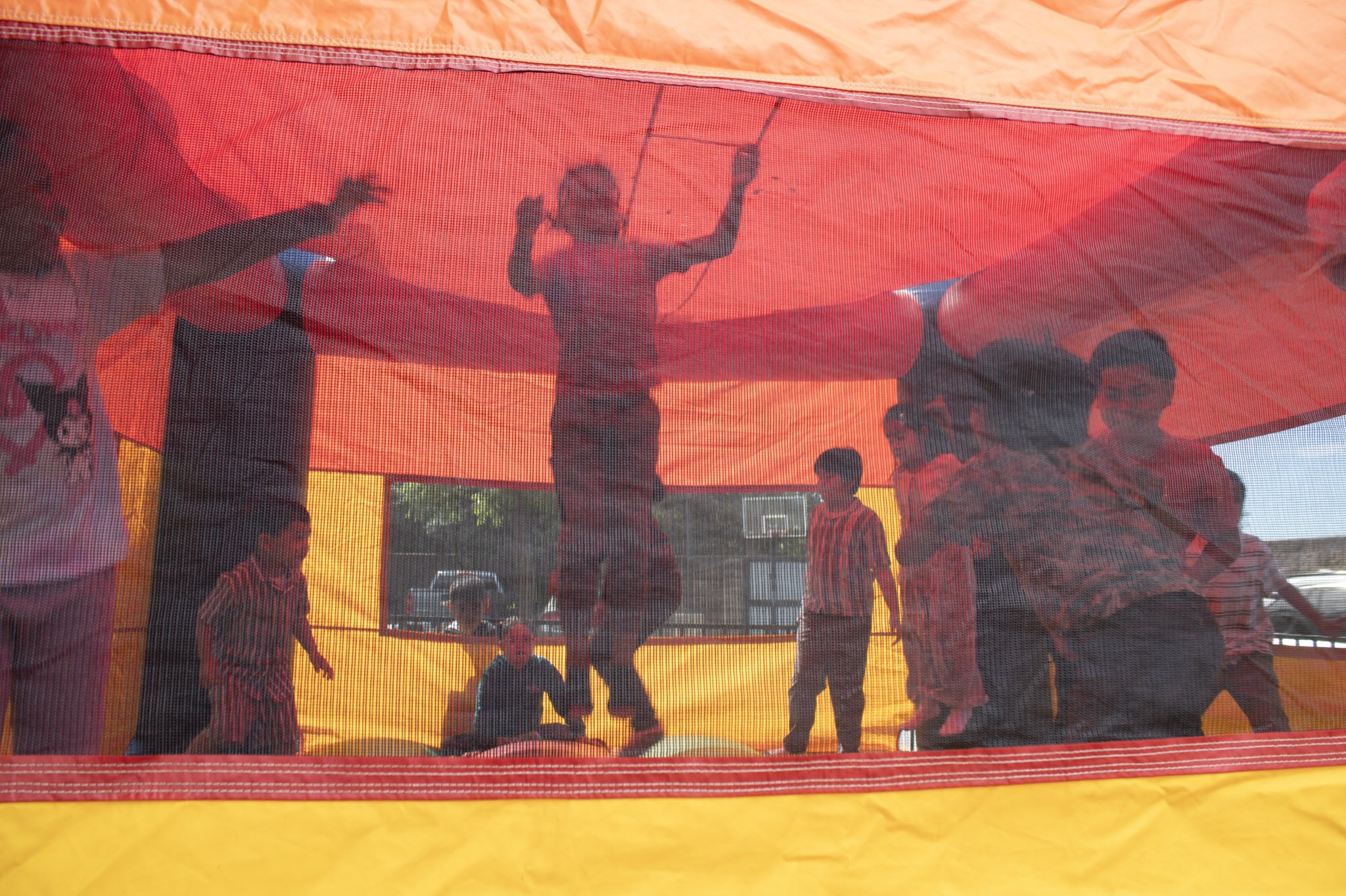 Children and adults playing inside a colorful bounce house with mesh sides, featuring a red and yellow color scheme, during daytime.