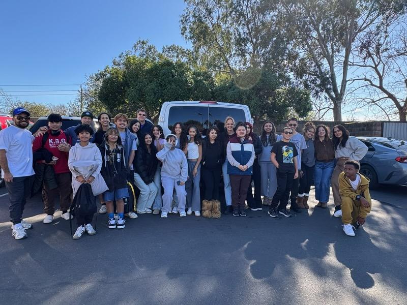 Group of diverse people, mostly children and teenagers, standing outdoors in front of a white van and trees, posing for a photo on a sunny day.