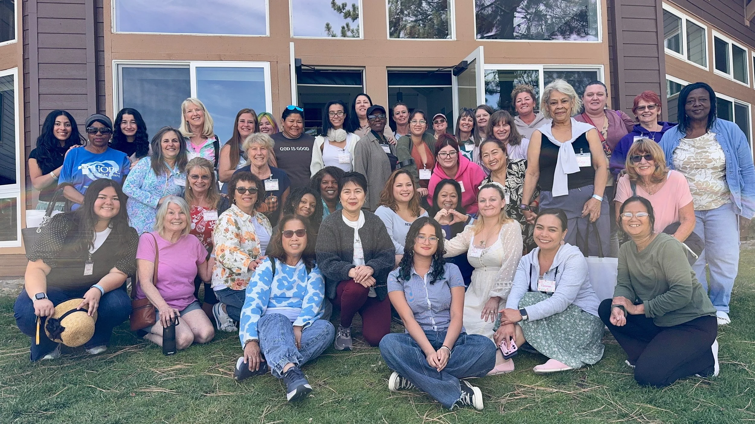Group of diverse women posed together outside in front of a building for a photo, some are sitting and some are standing, smiling and enjoying the moment.