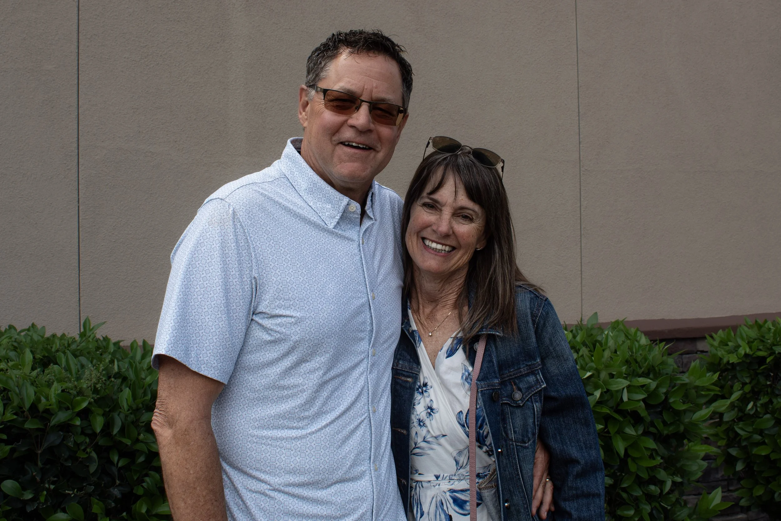 A man and woman smiling outdoors standing close together in front of green bushes and a beige wall, with all wearing casual attire including glasses.