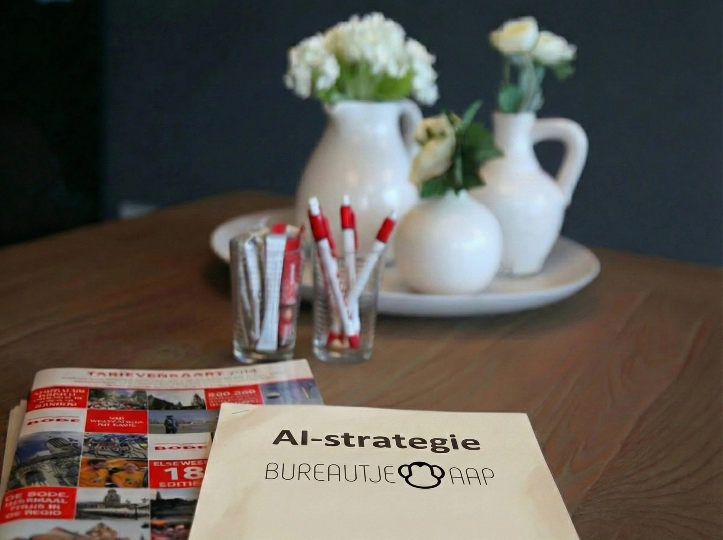Een tafel met een brochure en een document getiteld 'AI-strategie BUREAUTJE AAP', op de achtergrond drie witte vazen met bloemen, en een glas met rode en witte pennen.