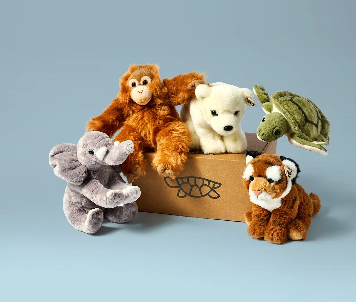 Verschillende knuffeldieren, waaronder een olifant, aap, hond, schildpad en tijger, naast een kartonnen doos met een leeuwentandlogo