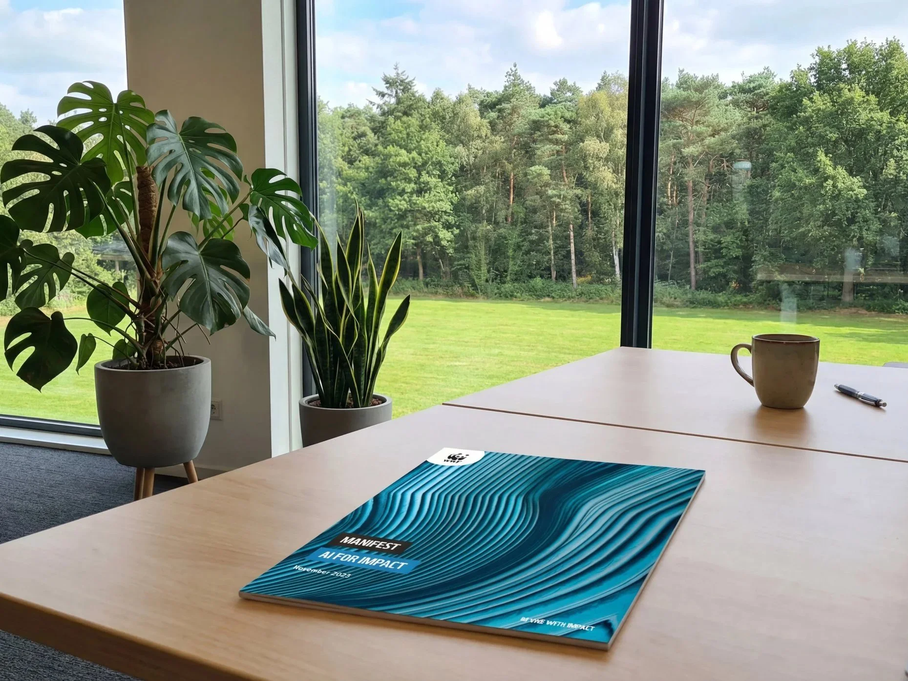 Een kantoorruimte met een tafel, twee planten in potten, een beker en een pen. Buiten is een groen landschap met bomen.