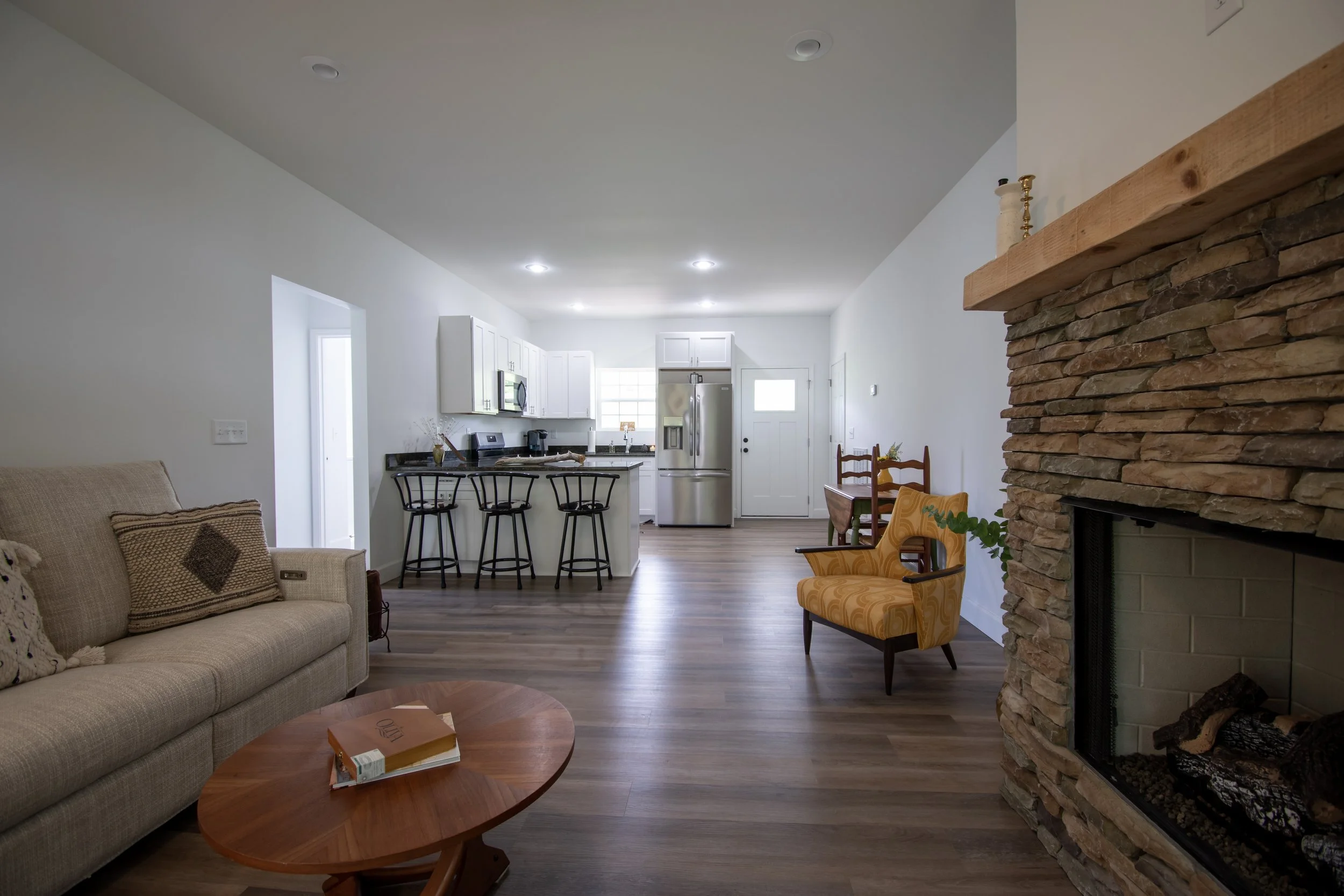 Spacious living room with beige sofa, wooden coffee table, yellow armchair, stone fireplace, and open kitchen with white cabinets and stainless steel appliances.
