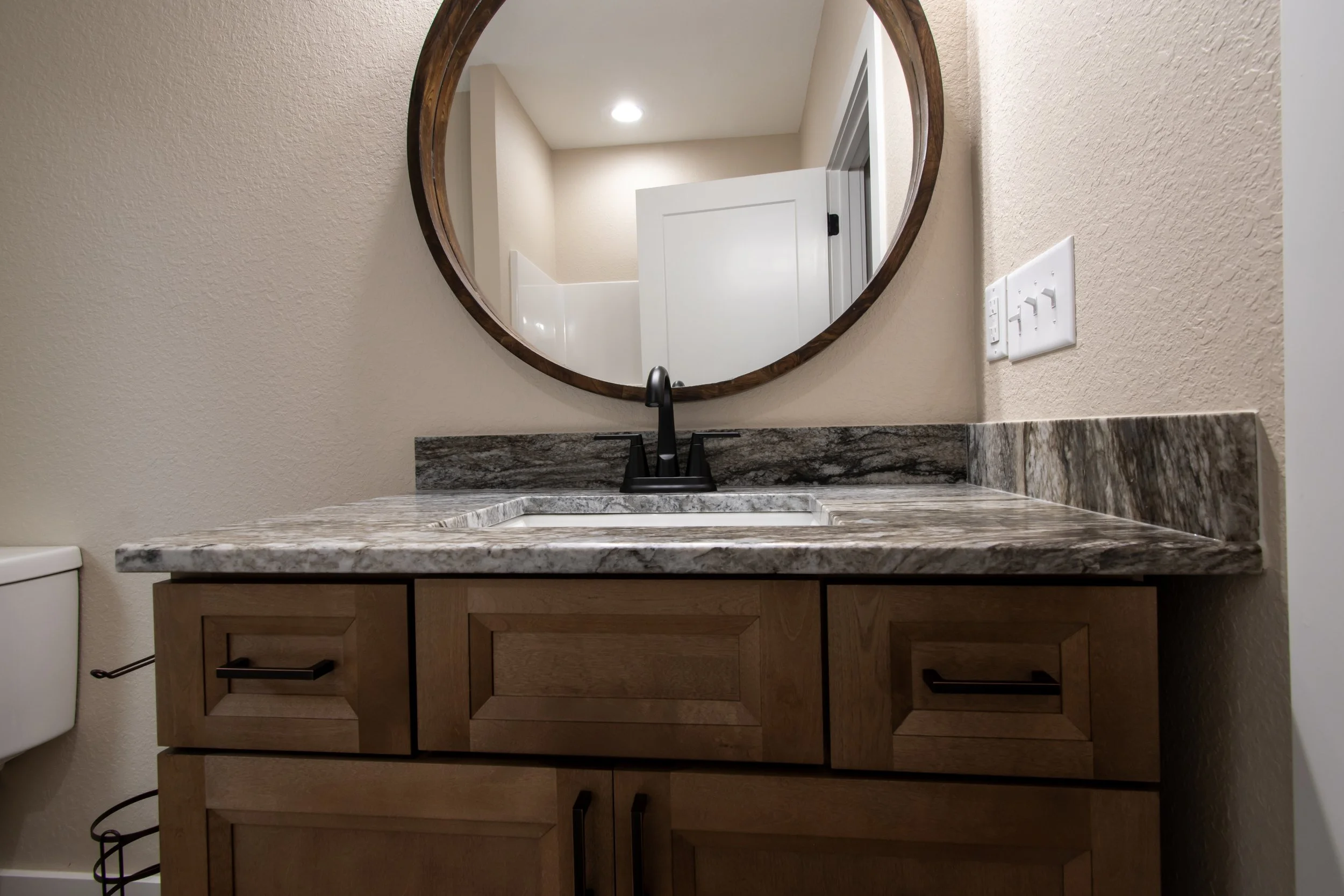 Bathroom vanity with a granite countertop, wooden cabinet, black faucet, and a round wooden mirror above. Part of a white toilet and a wall with a power outlet are visible.