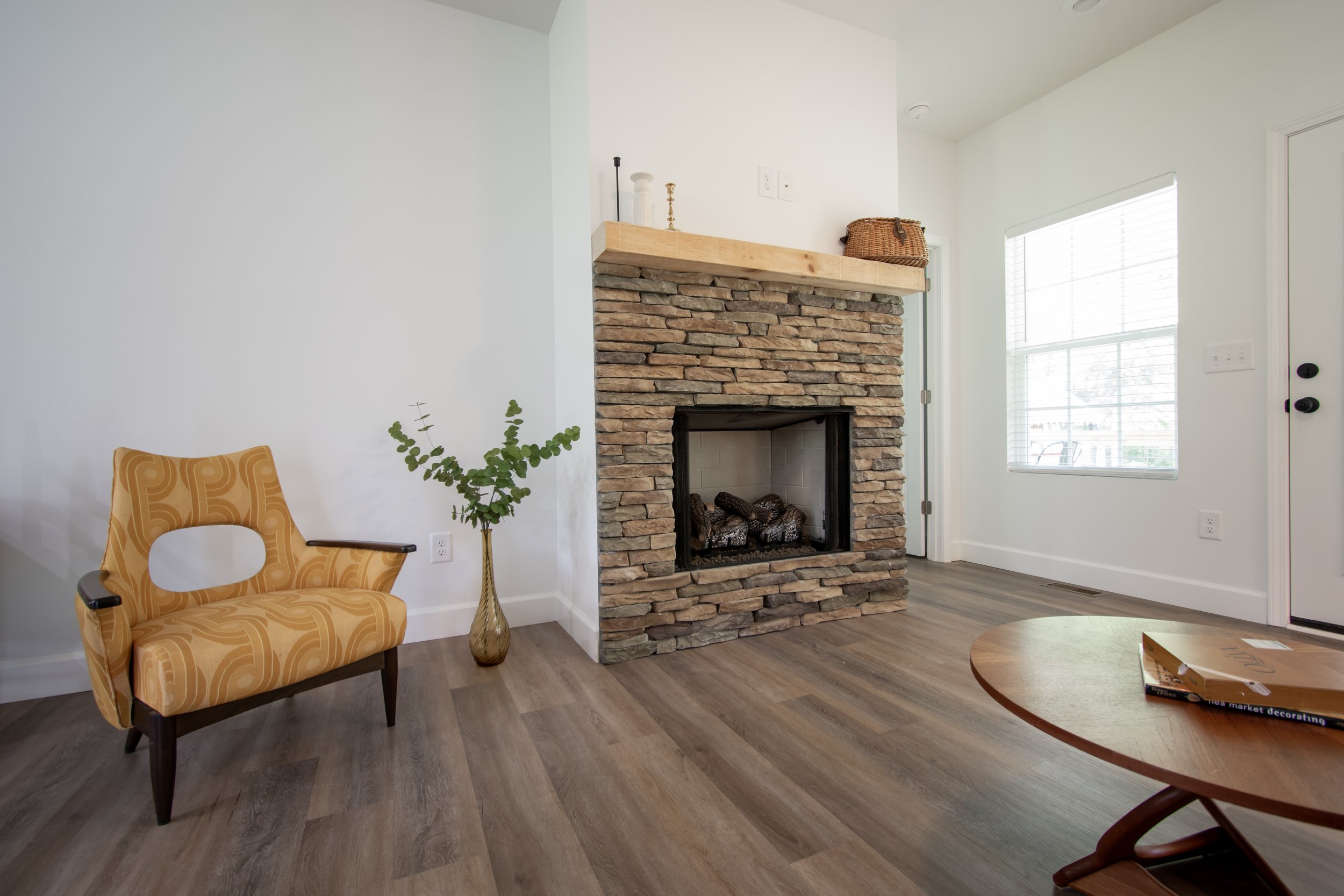Living room with a stone fireplace, a yellow patterned armchair, a small table with a tray, a vase with green foliage, and a window with blinds.
