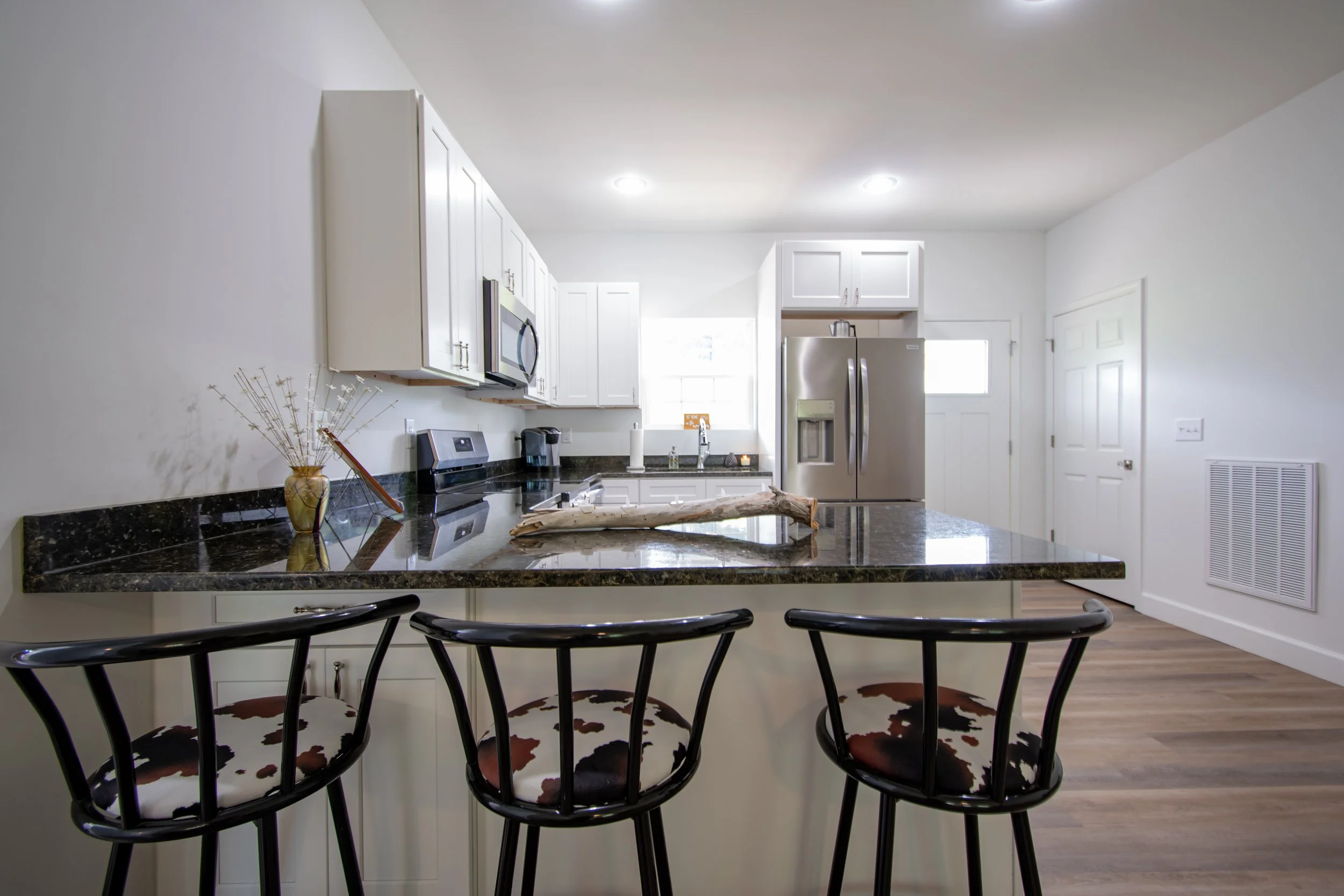 Modern kitchen with white cabinets, stainless steel appliances, black granite countertops, and three barstools with cow print cushions at a breakfast bar.