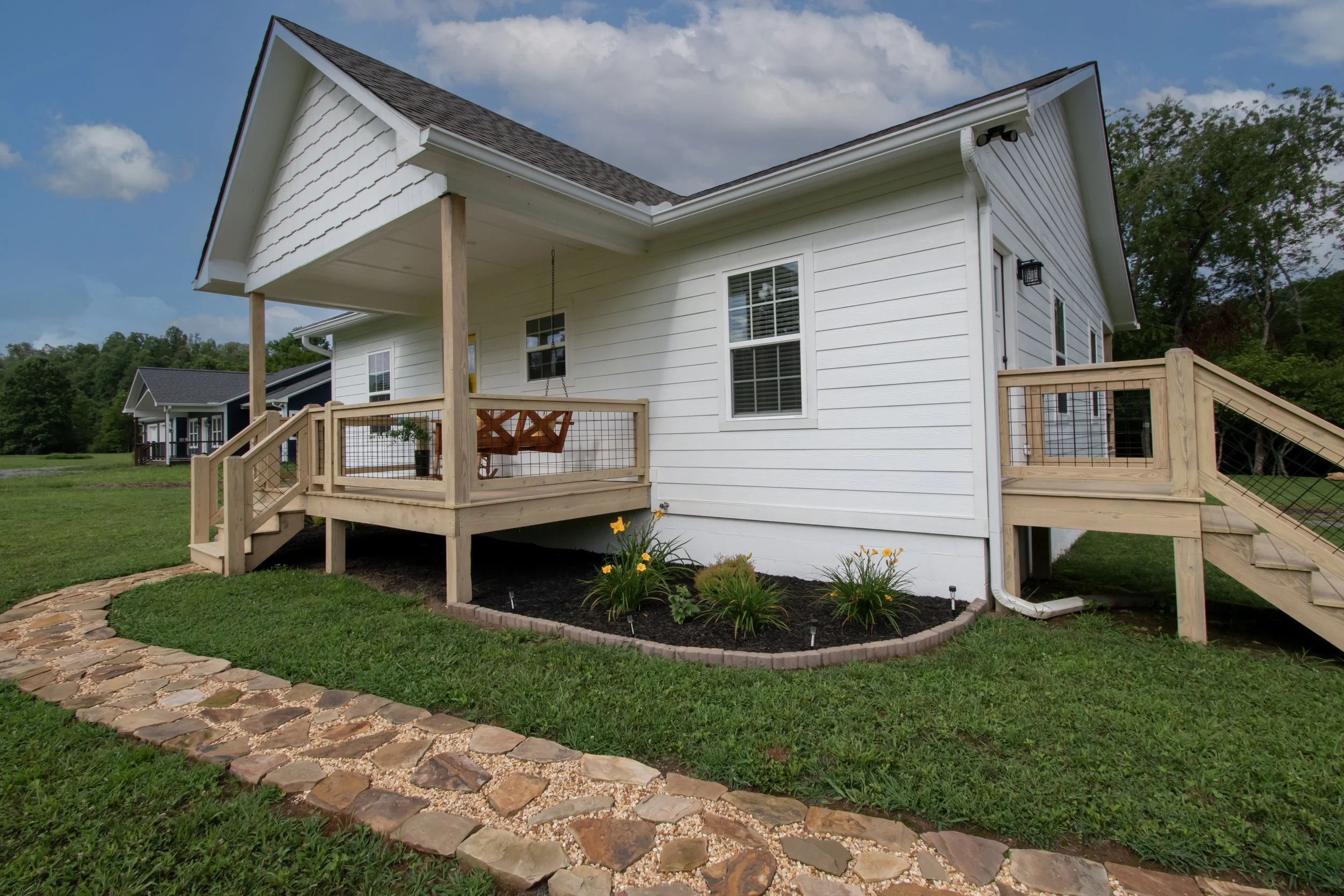 Newly built white house with a wooden deck and stairs, garden with flowers, grassy yard, and a stone pathway on a cloudy day.