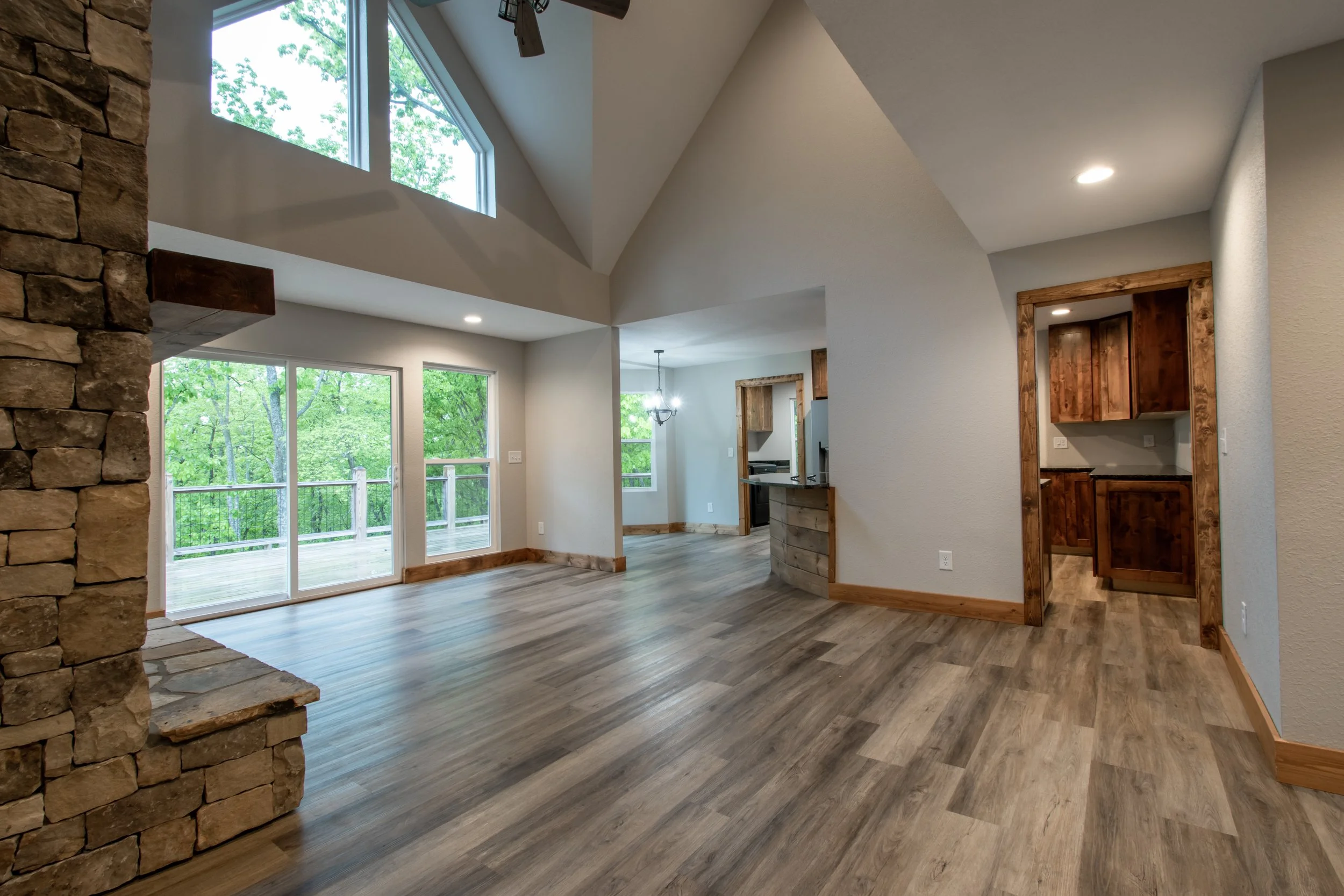 Empty living room with large windows, wooden floors, stone fireplace on the left, and kitchen with wooden cabinets visible through an open doorway.