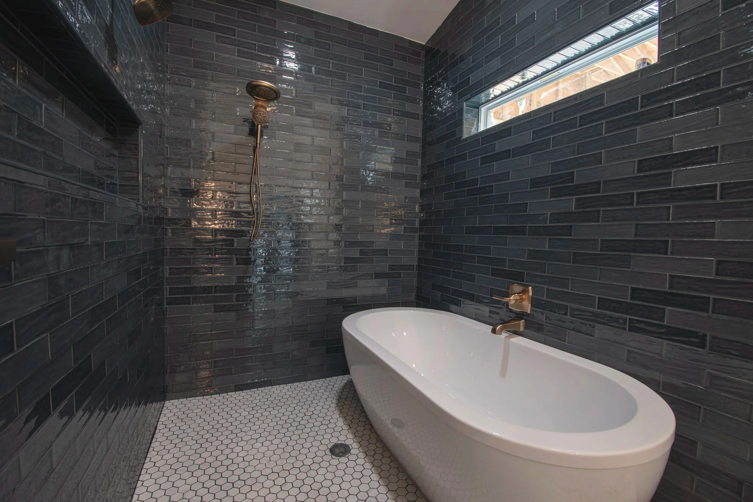 Modern bathroom with black tiled walls, a white freestanding bathtub, a gold faucet, a wall-mounted shower head, and a small horizontal window.
