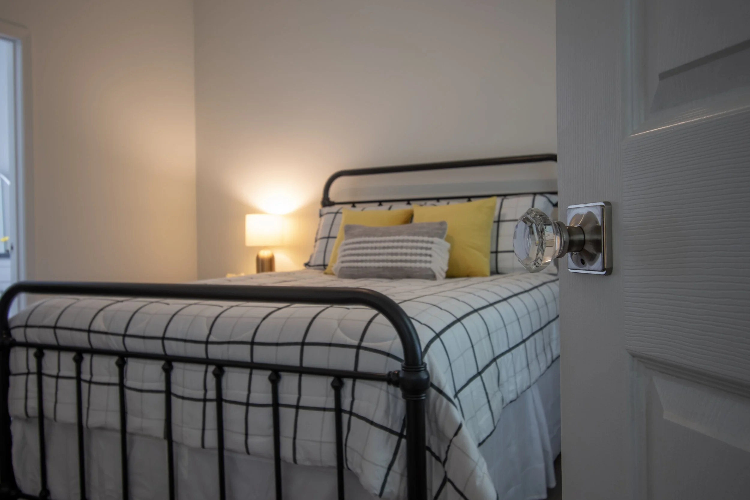 A neatly made bed with a black metal frame, striped black and white bedding, and yellow and gray pillows, in a room with a lit lamp on a nightstand.