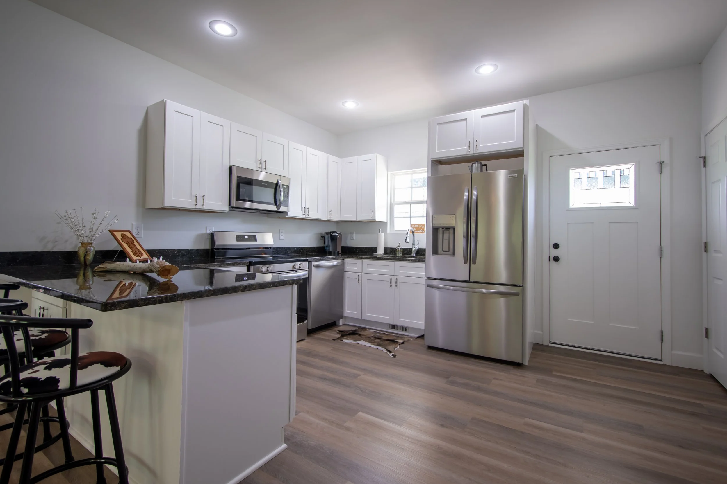 Modern kitchen with white cabinets, stainless steel appliances, black granite countertops, a small window, and wooden flooring.