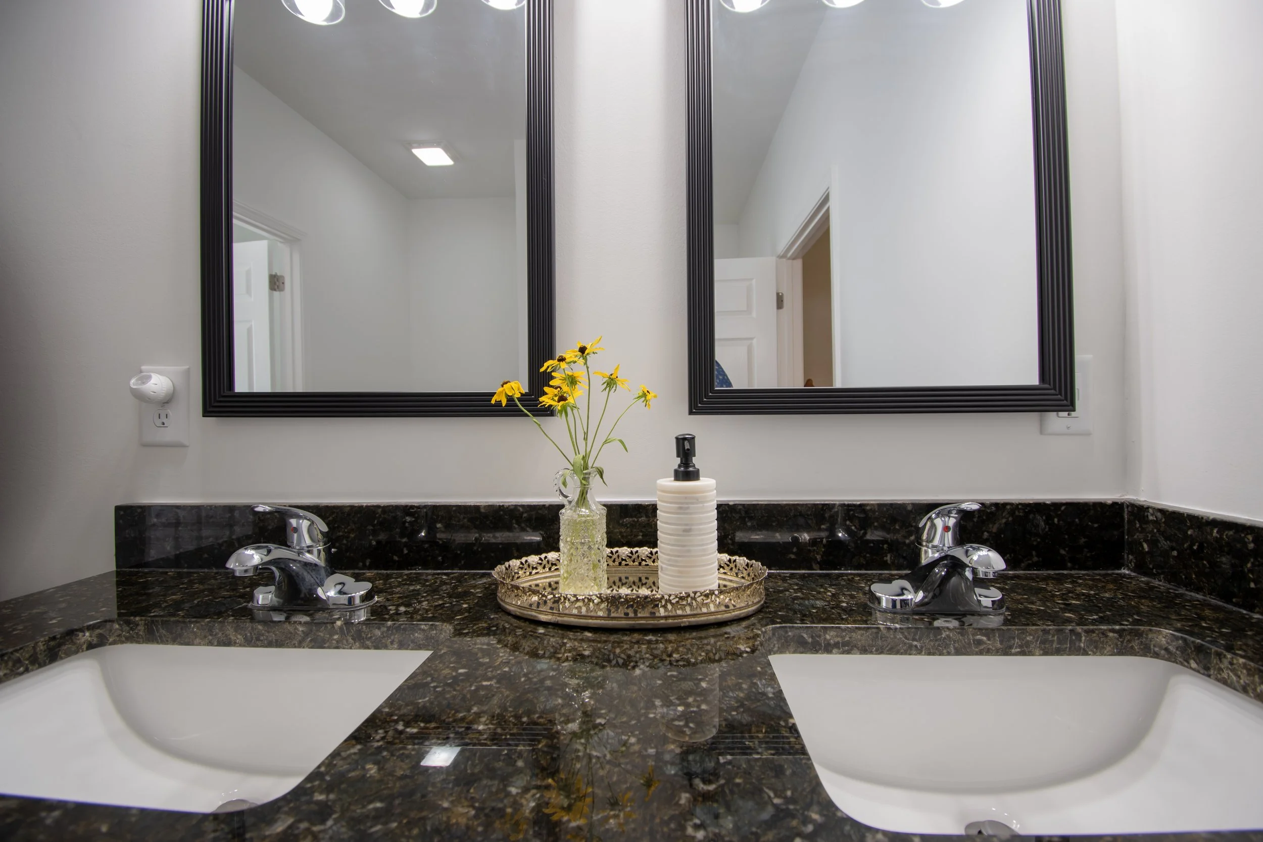 A double bathroom vanity with a dark granite countertop, two white sinks, and two mirrors. In the center, on a decorative tray, there is a vase with yellow flowers and a soap dispenser.