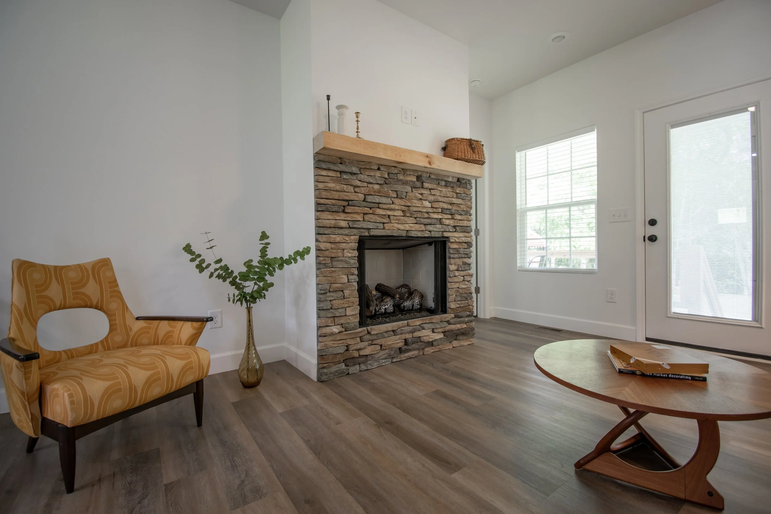 Living room with a stone fireplace, a yellow patterned armchair, a glass vase with greenery, a wooden coffee table with books, and a large window and glass door allowing natural light.