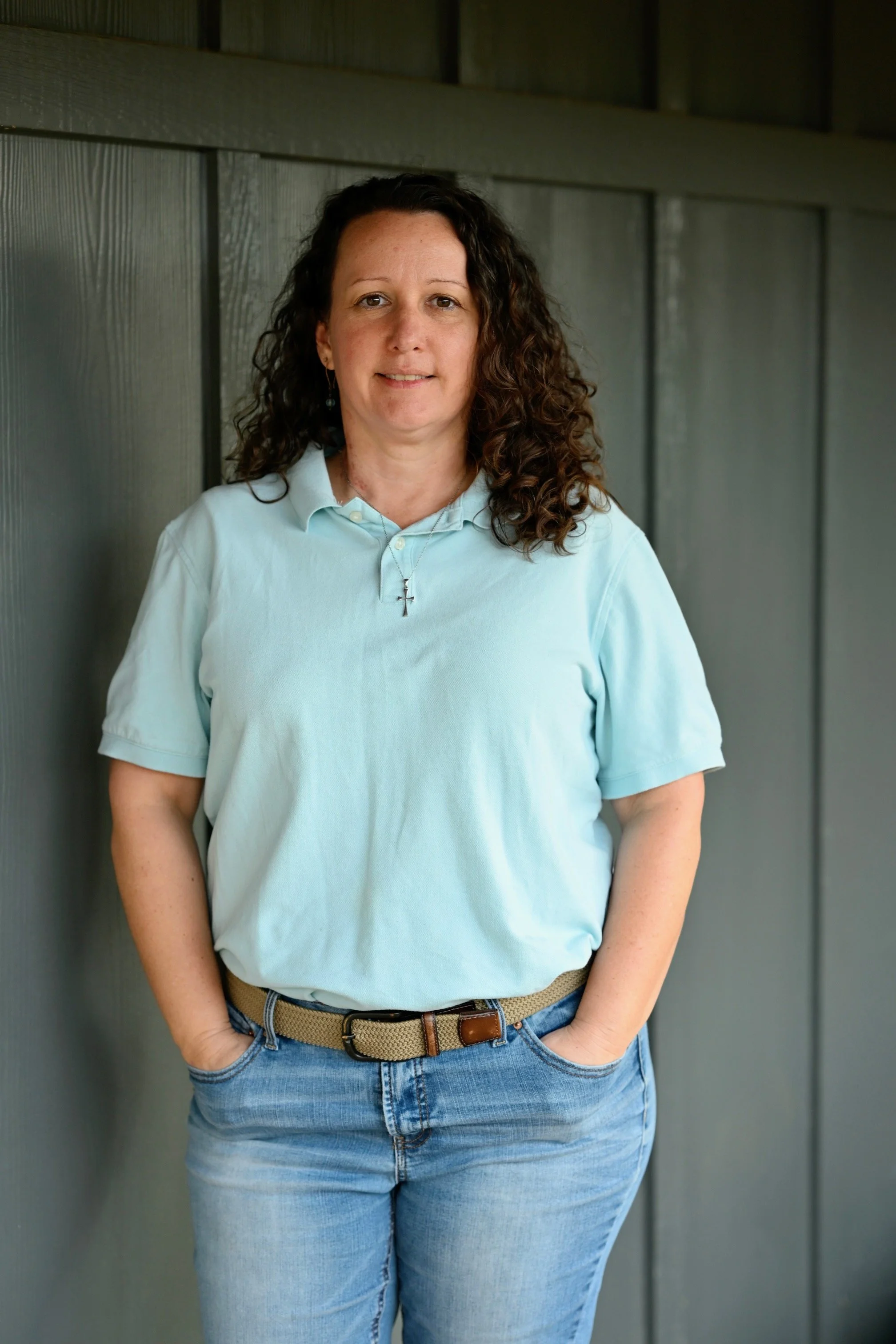 A woman with curly brown hair, wearing a light blue polo shirt and jeans, standing against a gray wooden wall. She has her hands in her pockets and is looking at the camera.