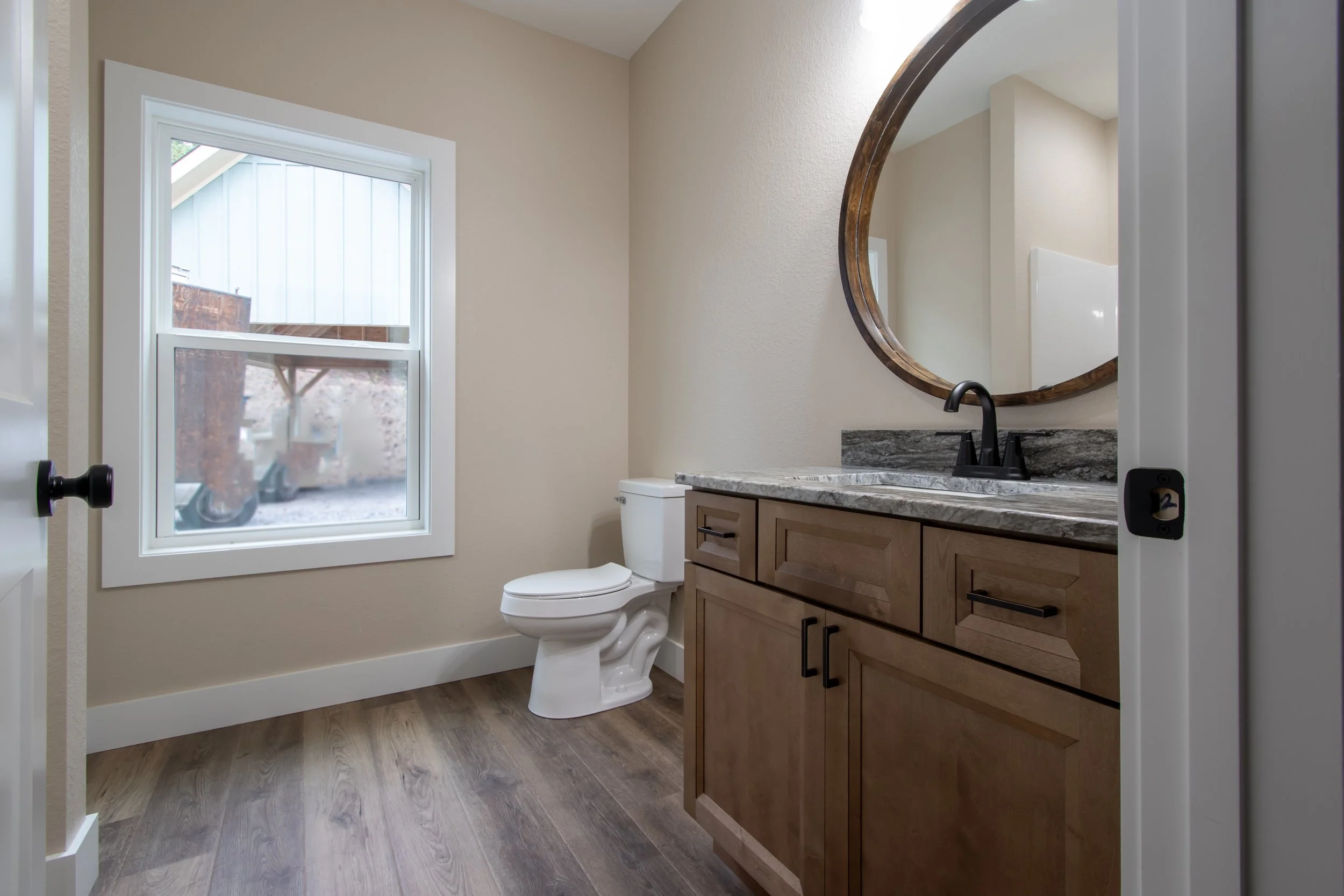 A small bathroom with a beige wall, a white toilet, a wooden cabinet with a granite countertop, a round wooden-framed mirror, and a window showing some outdoor elements.