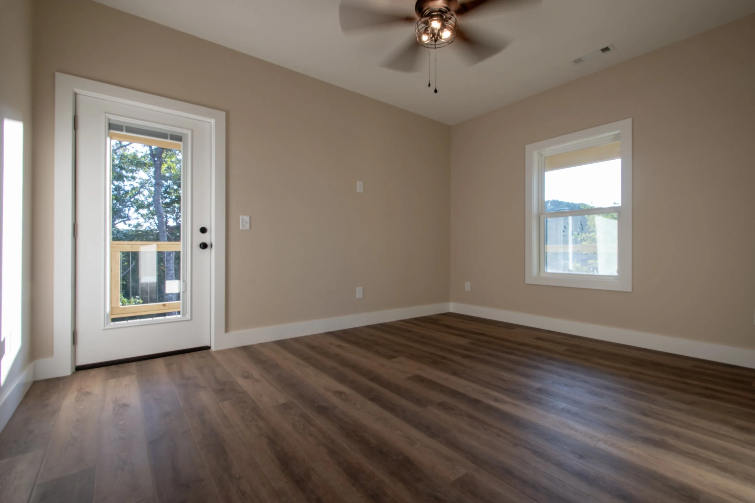 Empty room with hardwood flooring, beige walls, two windows, a glass door with a wooden railing, and a ceiling fan.