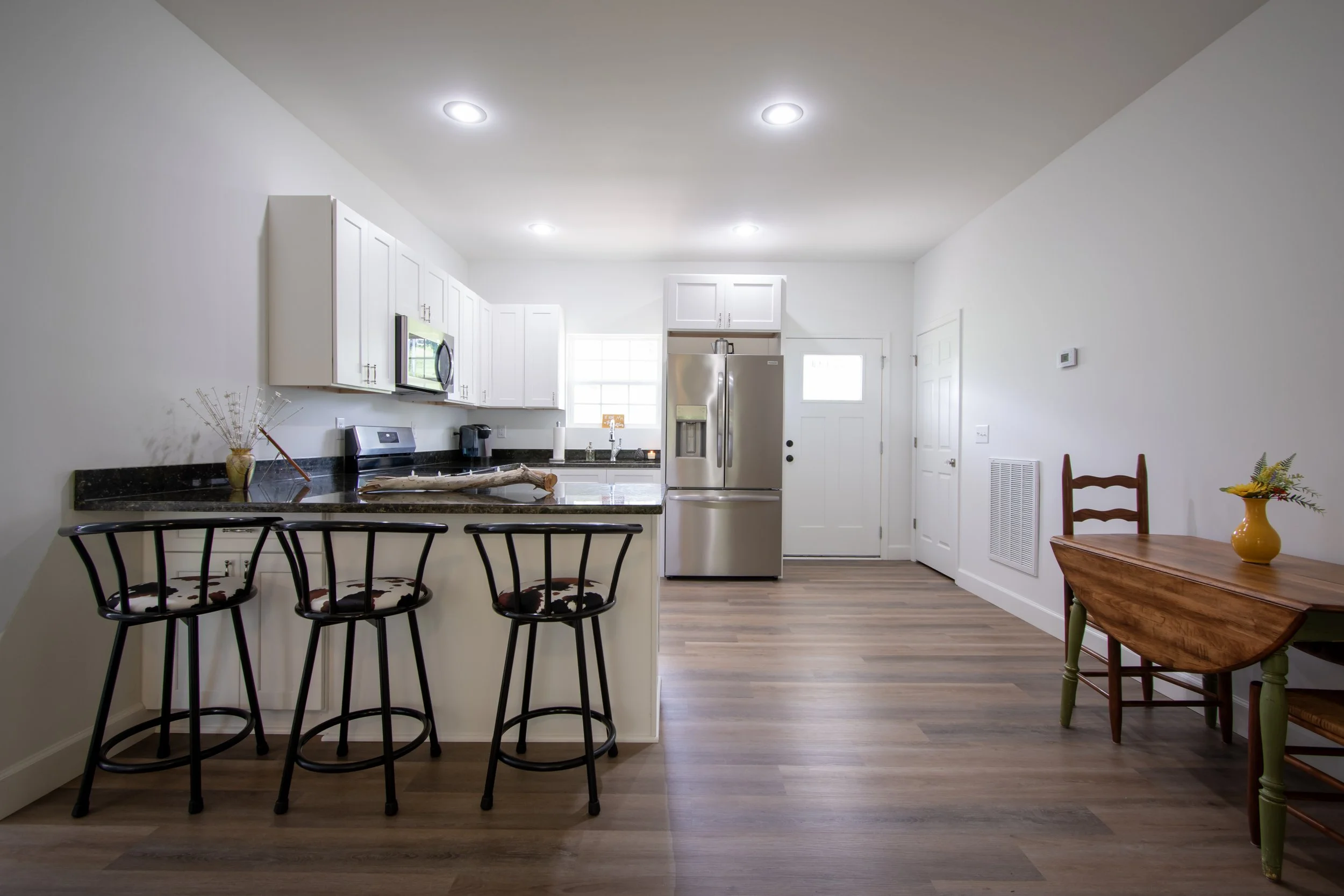 Bright kitchen with white cabinets, stainless steel refrigerator, black granite countertops, and a breakfast bar with three black chairs.