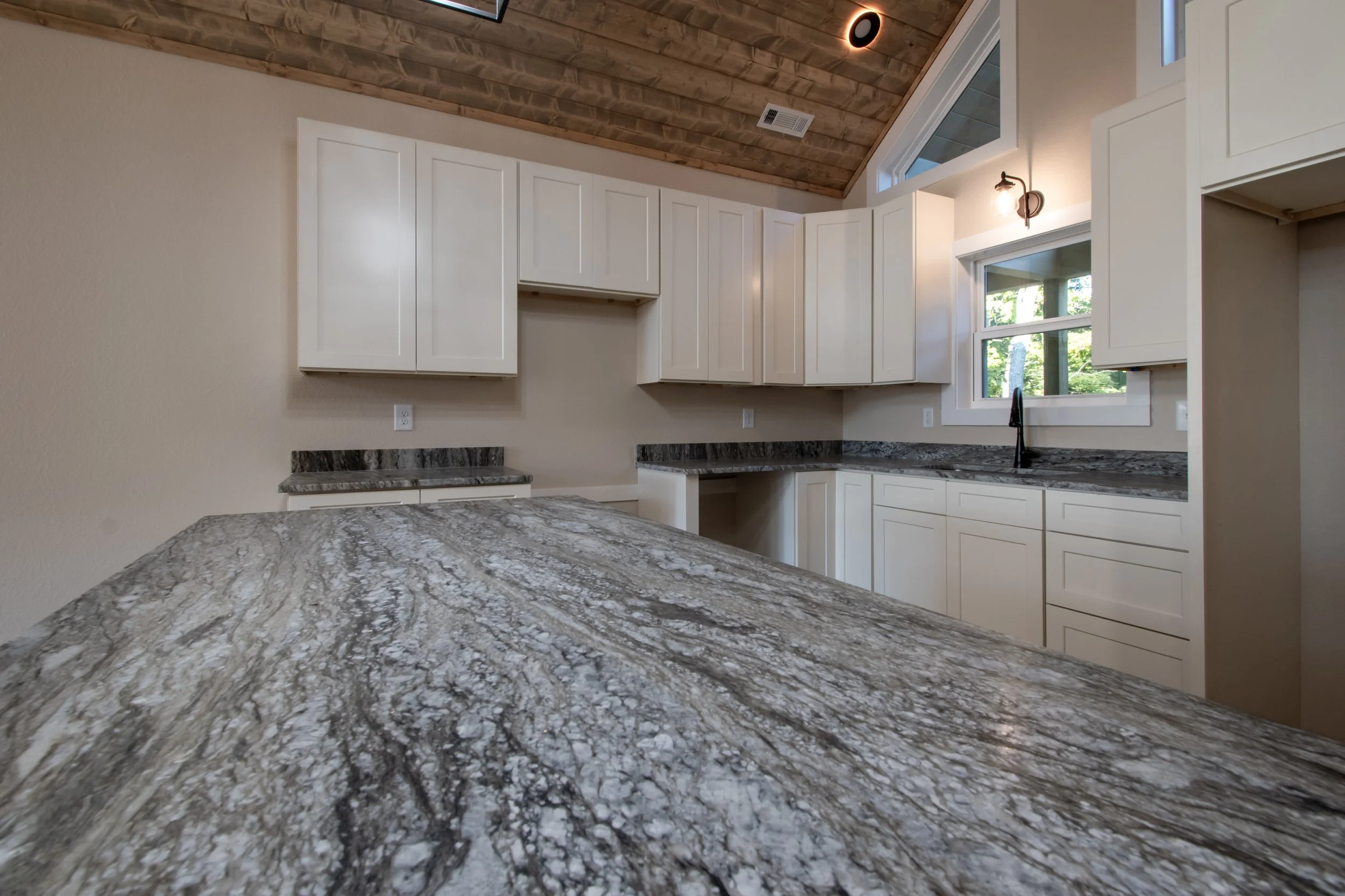 Empty kitchen with white cabinets, granite countertops, black faucet, and wooden vaulted ceiling.