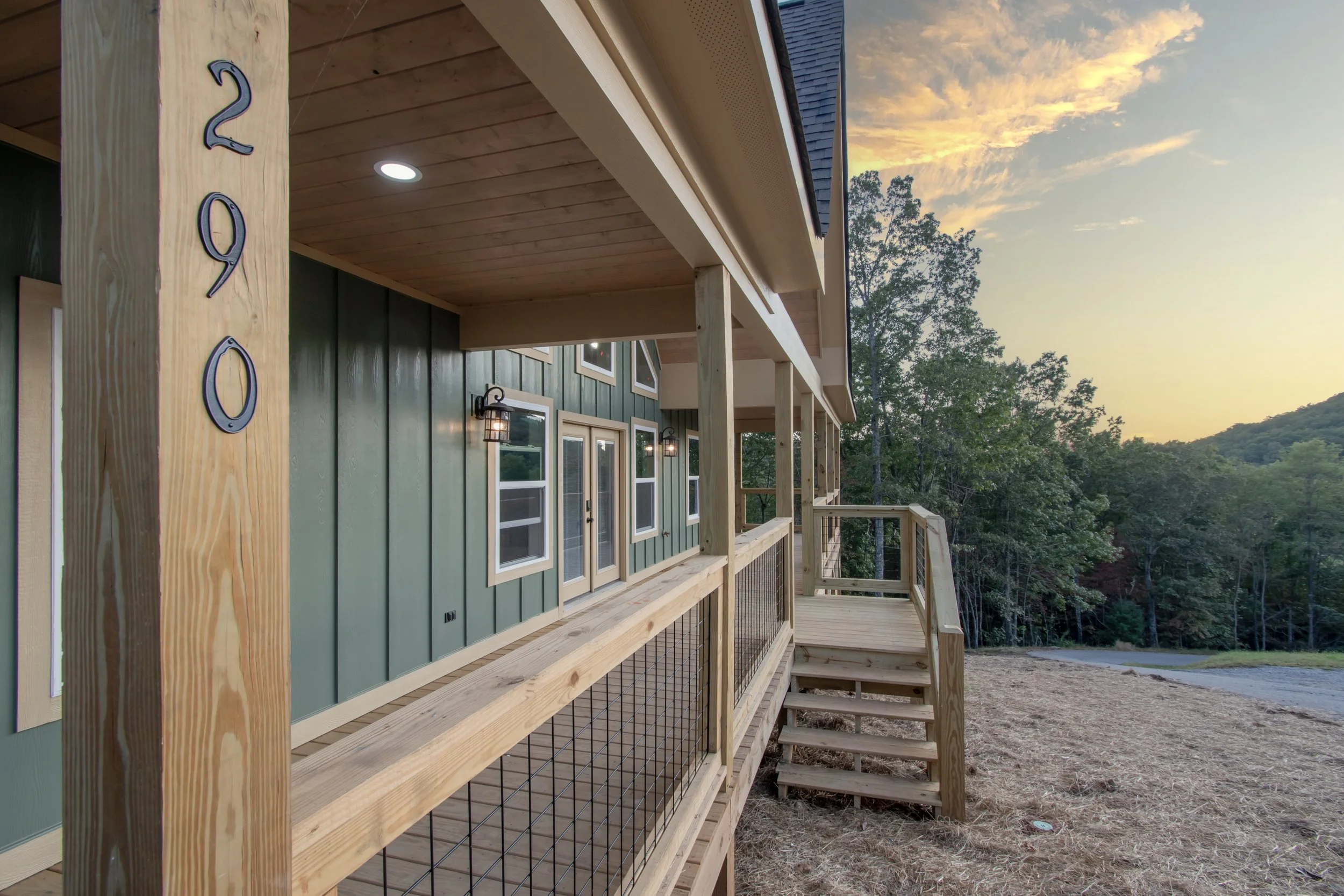 Newly constructed green house with a wooden deck, stairs, and railing, set against a scenic countryside sunset with trees and distant hills.