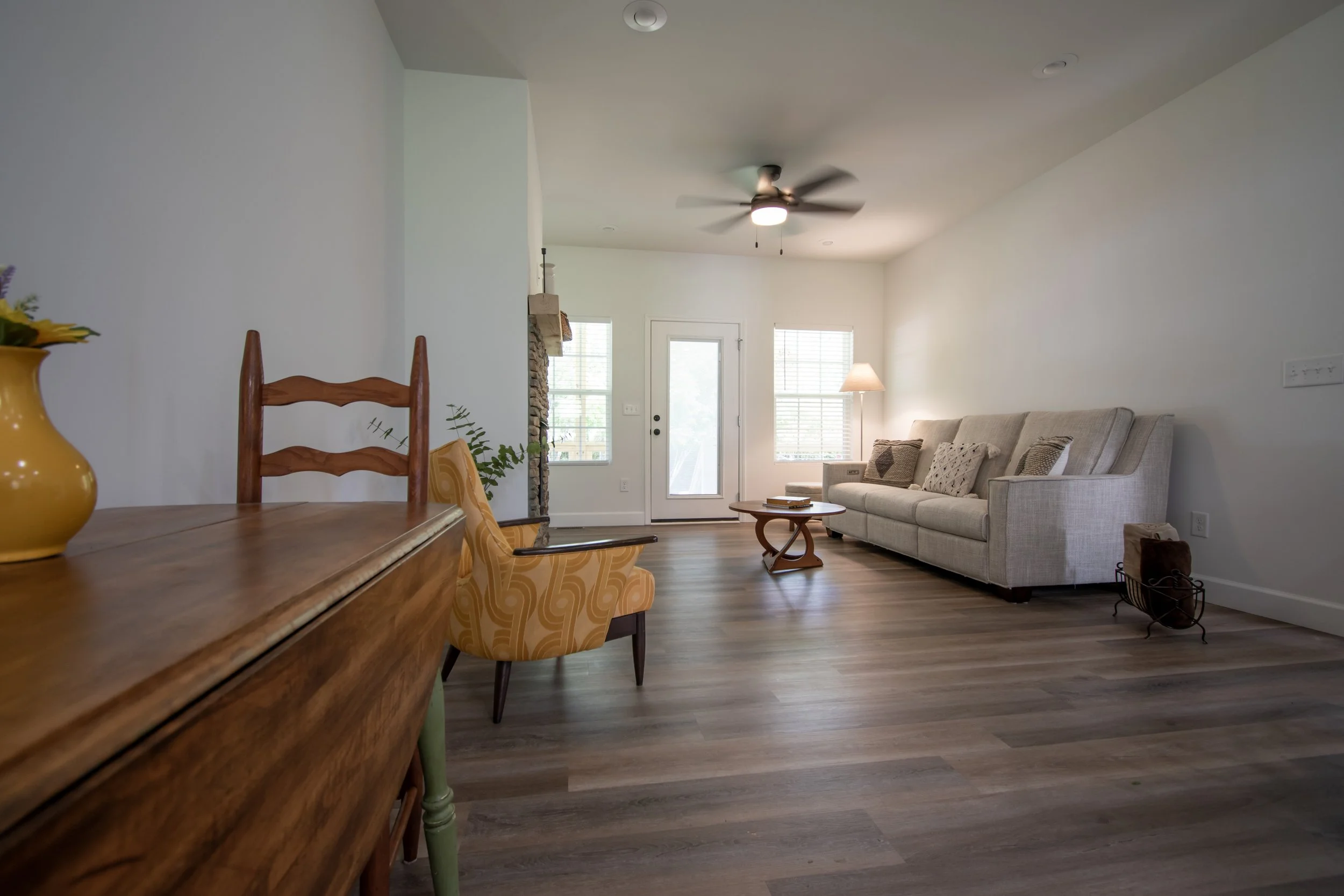 Living room with beige sofa, wooden coffee table, floor lamp, ceiling fan, and wooden dining table with chairs, hardwood floors, and doors and windows with blinds.
