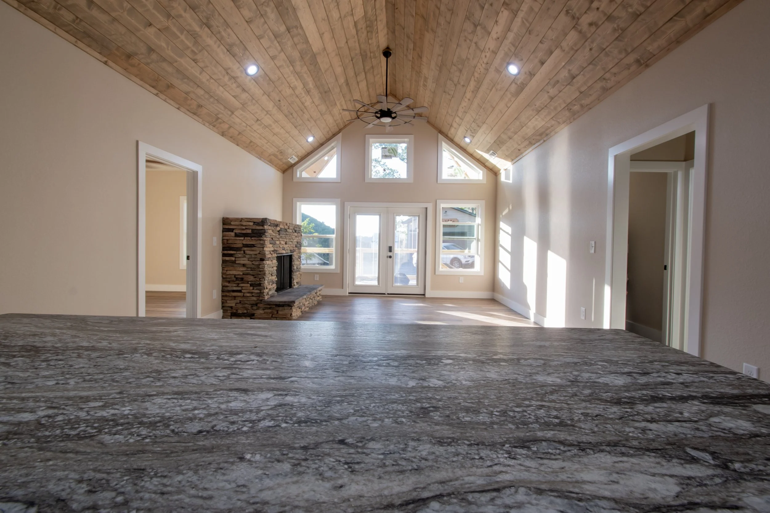 Empty living room with vaulted wooden ceiling, large windows, a stone fireplace, and a door leading outside, viewed from a countertop in the foreground.