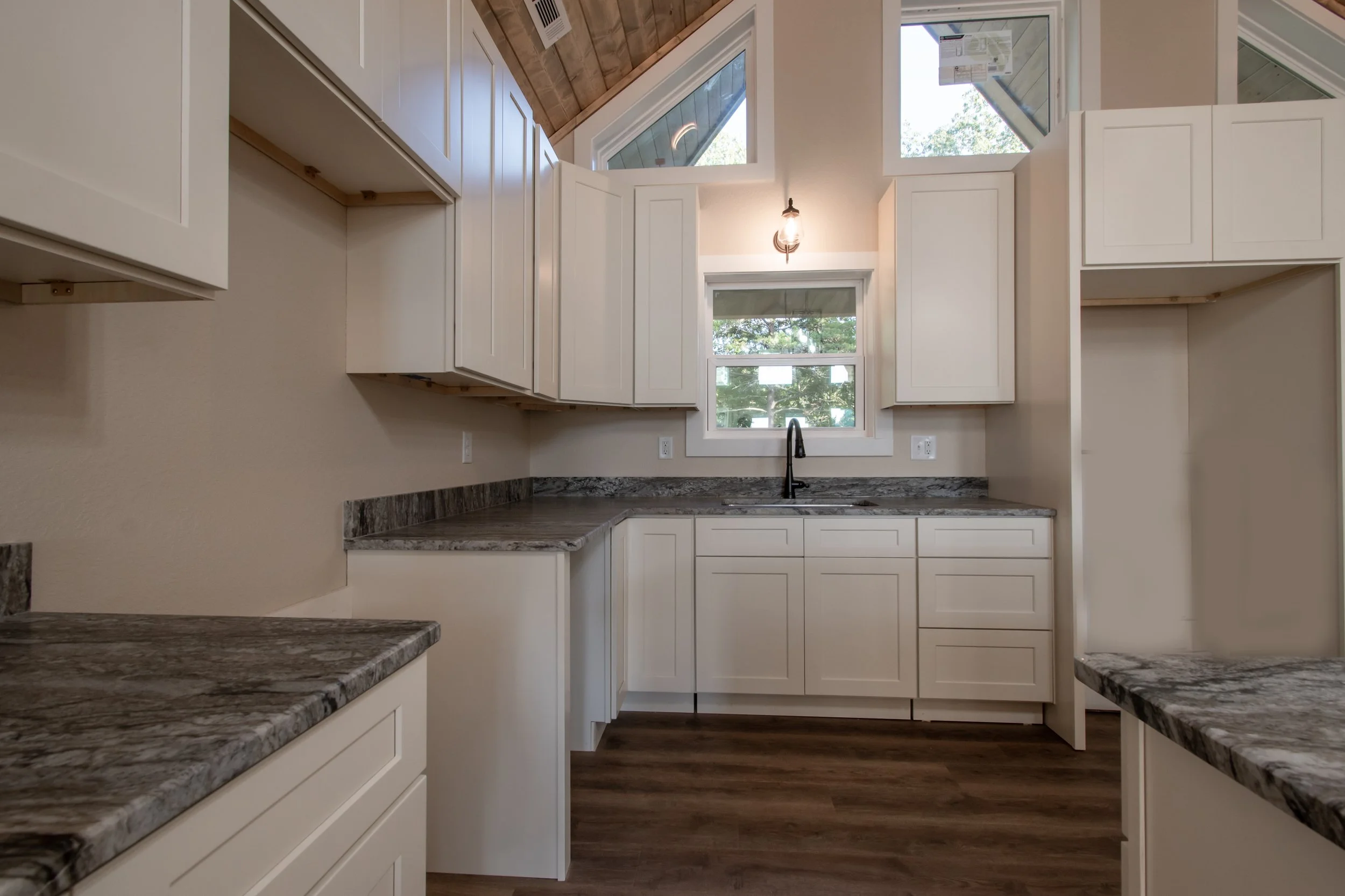 Modern kitchen with white cabinets, granite countertops, and a window above the sink, with a sloped wooden ceiling and hardwood flooring.