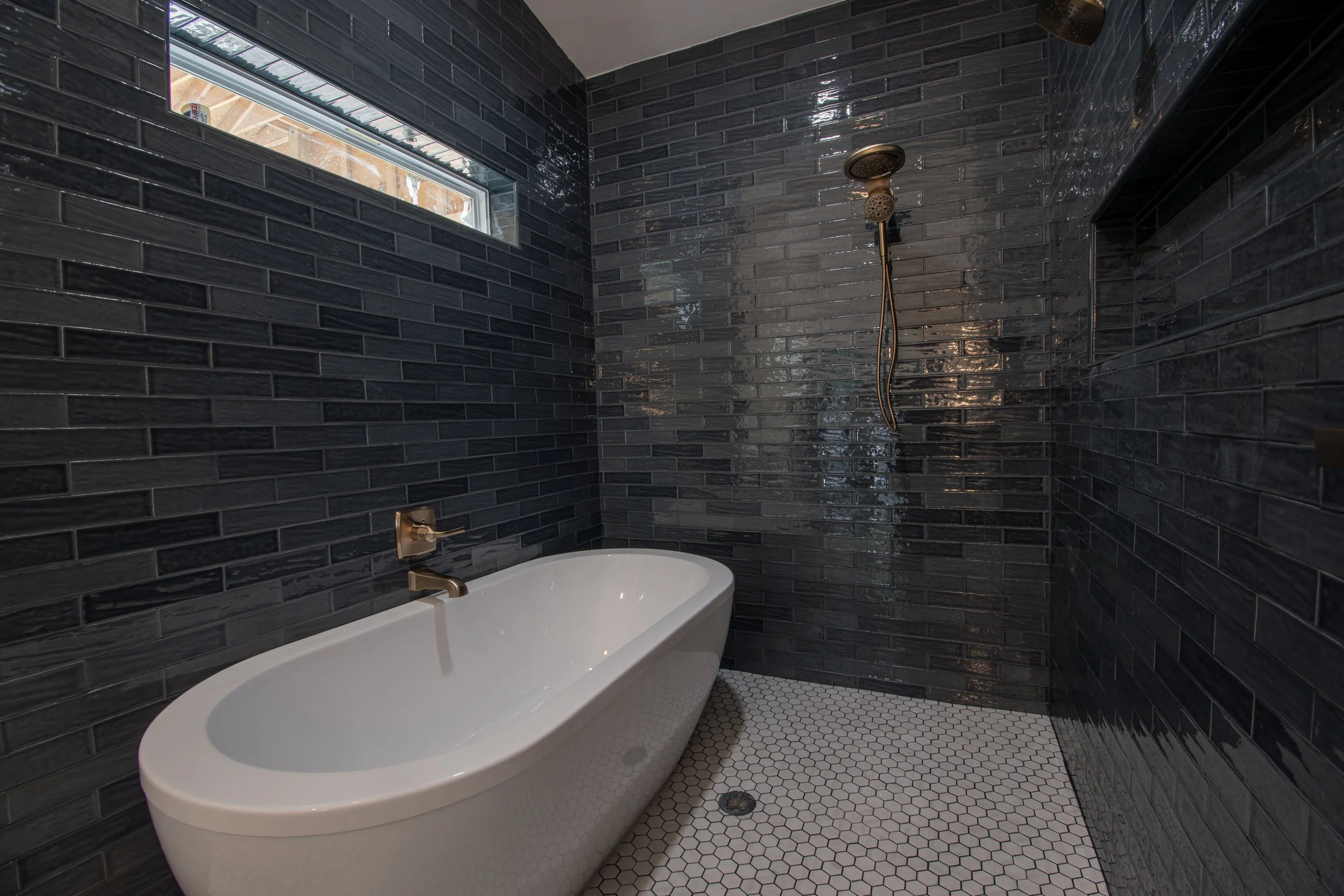Luxury bathroom with black tiled walls, white bathtub, small white hexagonal floor tiles, and a small window.
