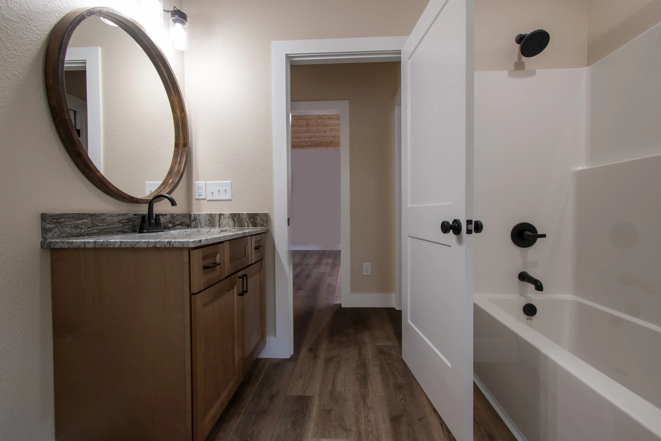 Bathroom with wooden vanity, marble countertop, oval mirror, and a bathtub with black fixtures, and a doorway leading to a room with hardwood flooring.