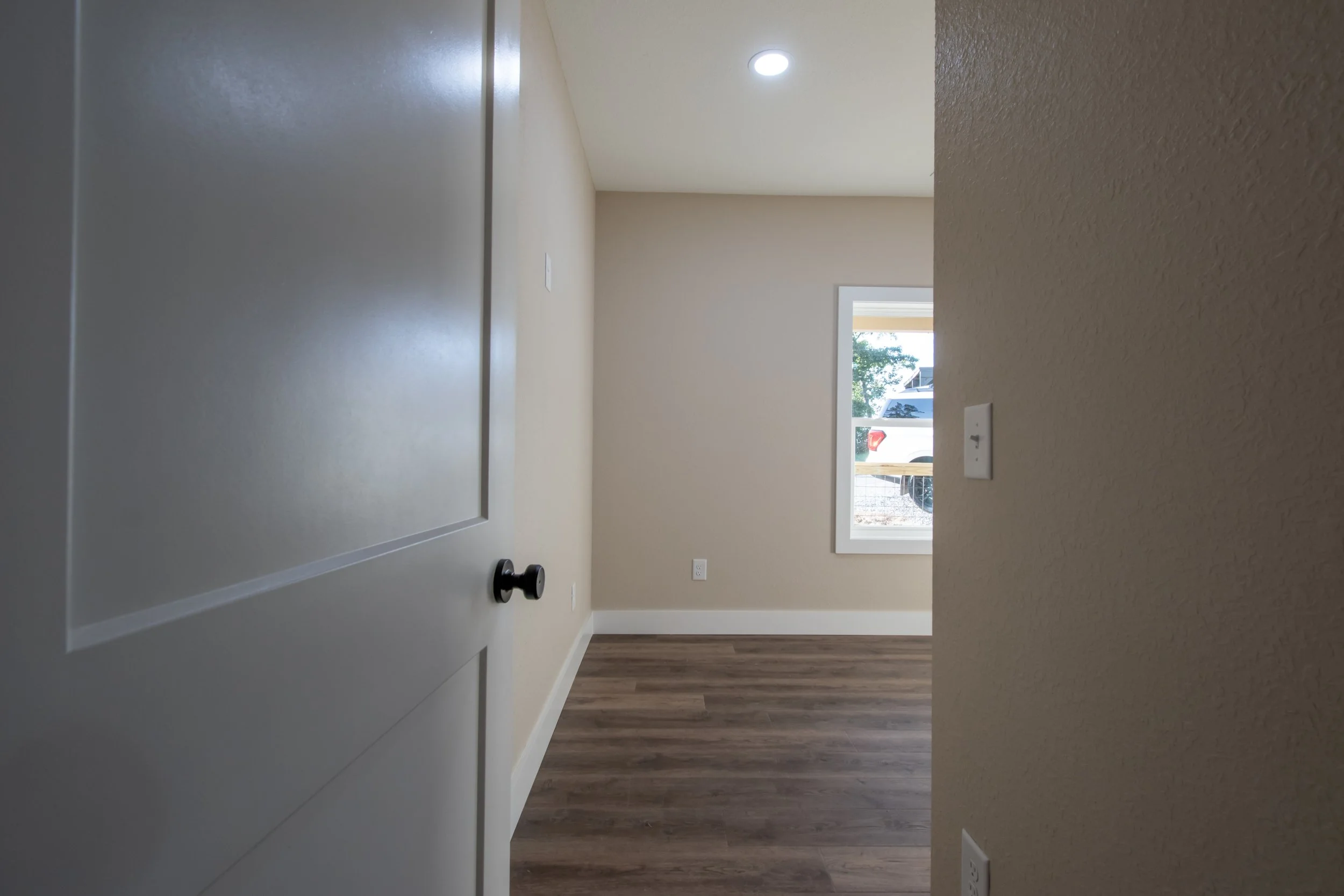Empty room with beige walls, wooden flooring, white baseboards, a window showing outdoor scenery, and a ceiling light.