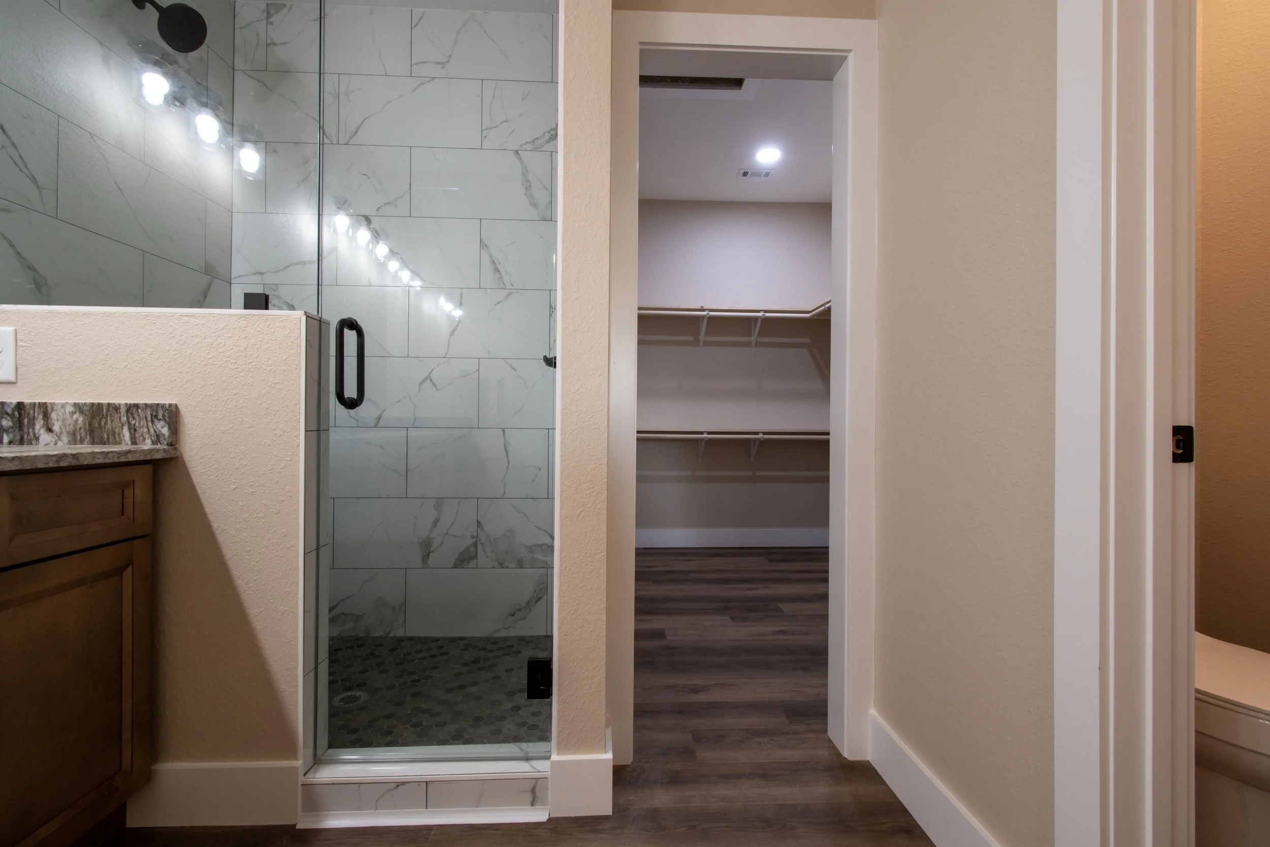 Bathroom with walk-in shower with marble tiles, wood vanity with granite countertop, and a walk-in closet with shelves and hanging rods.