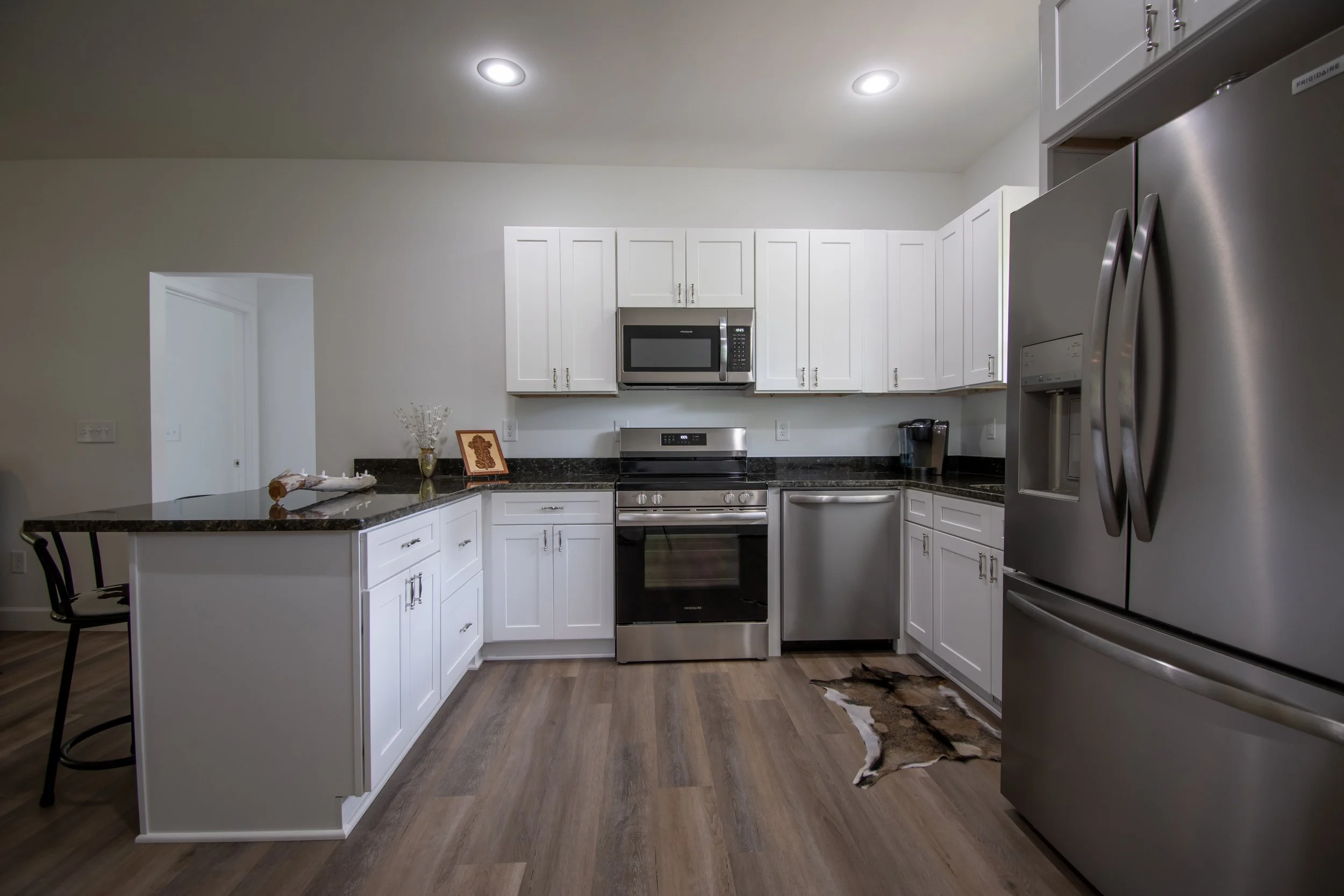 Modern kitchen with white cabinets, stainless steel appliances including a refrigerator, stove, microwave, and dishwasher. The floor is wood with a small animal hide rug near the corner.